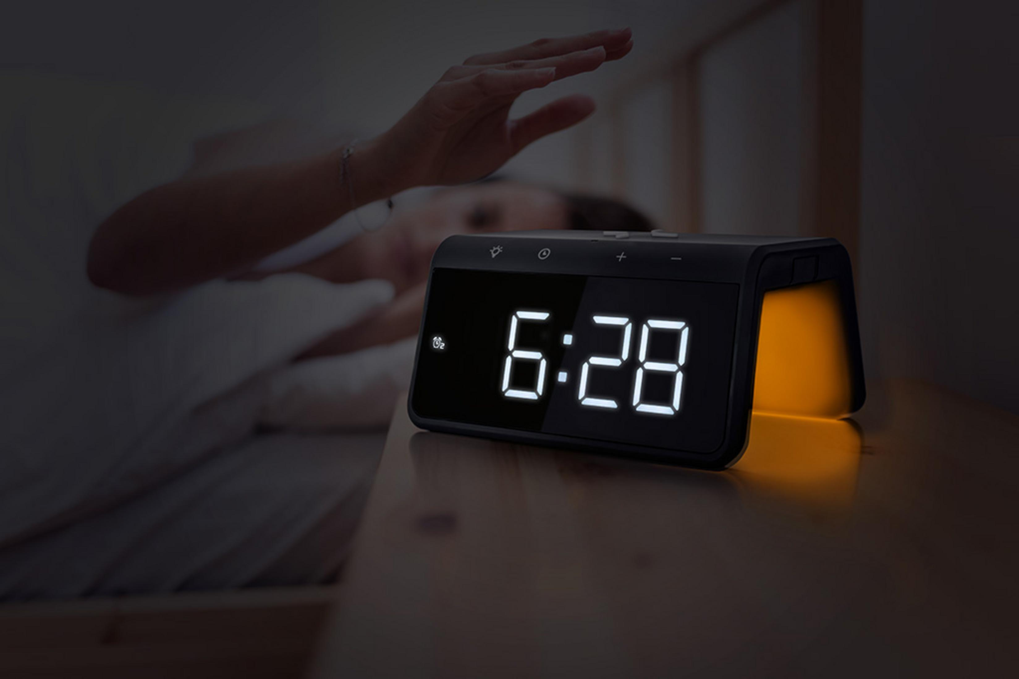A person reaches for a digital alarm clock displaying 6:28.