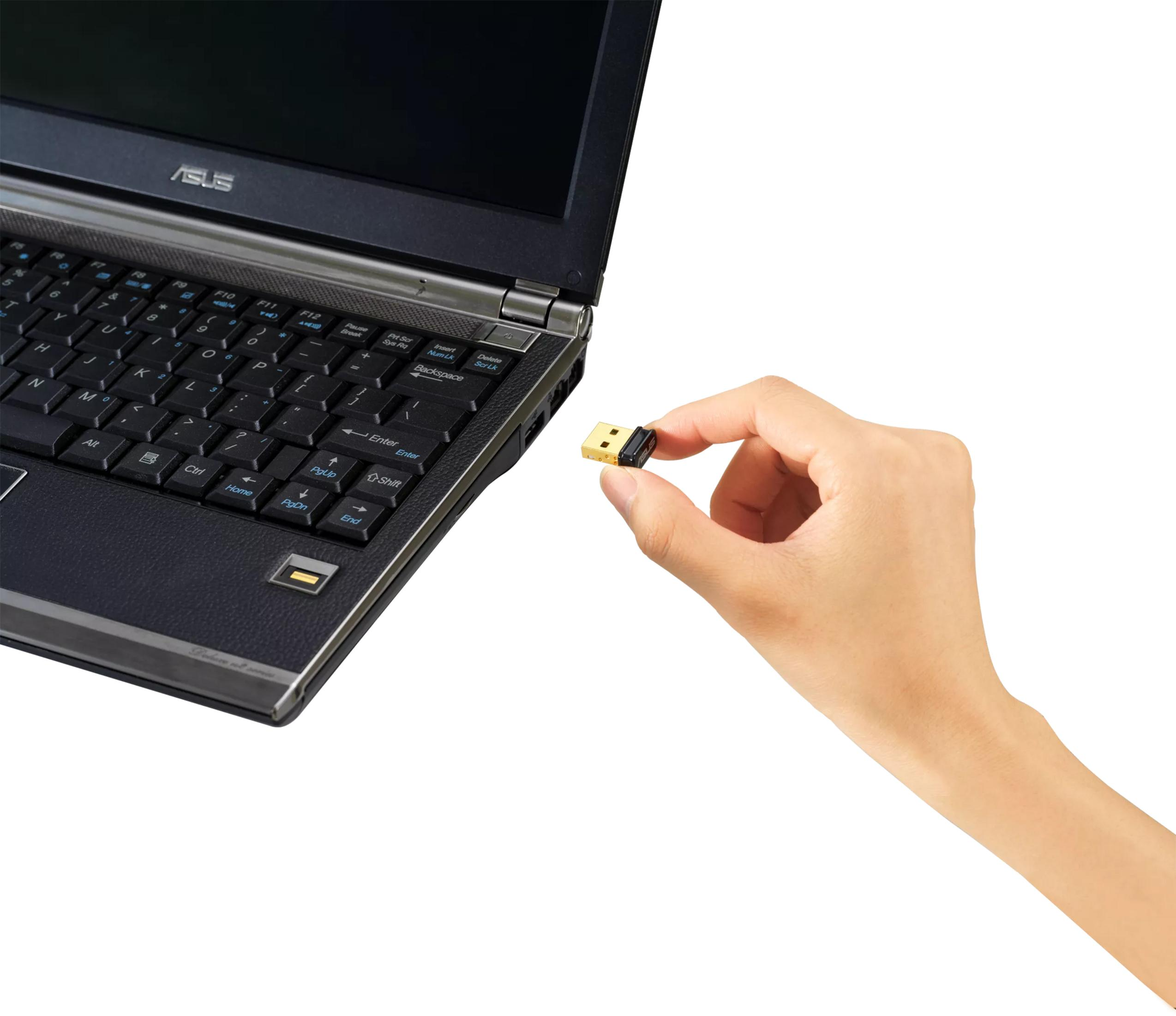 Una mano sostiene un pequeño receptor USB dorado cerca de un portátil.