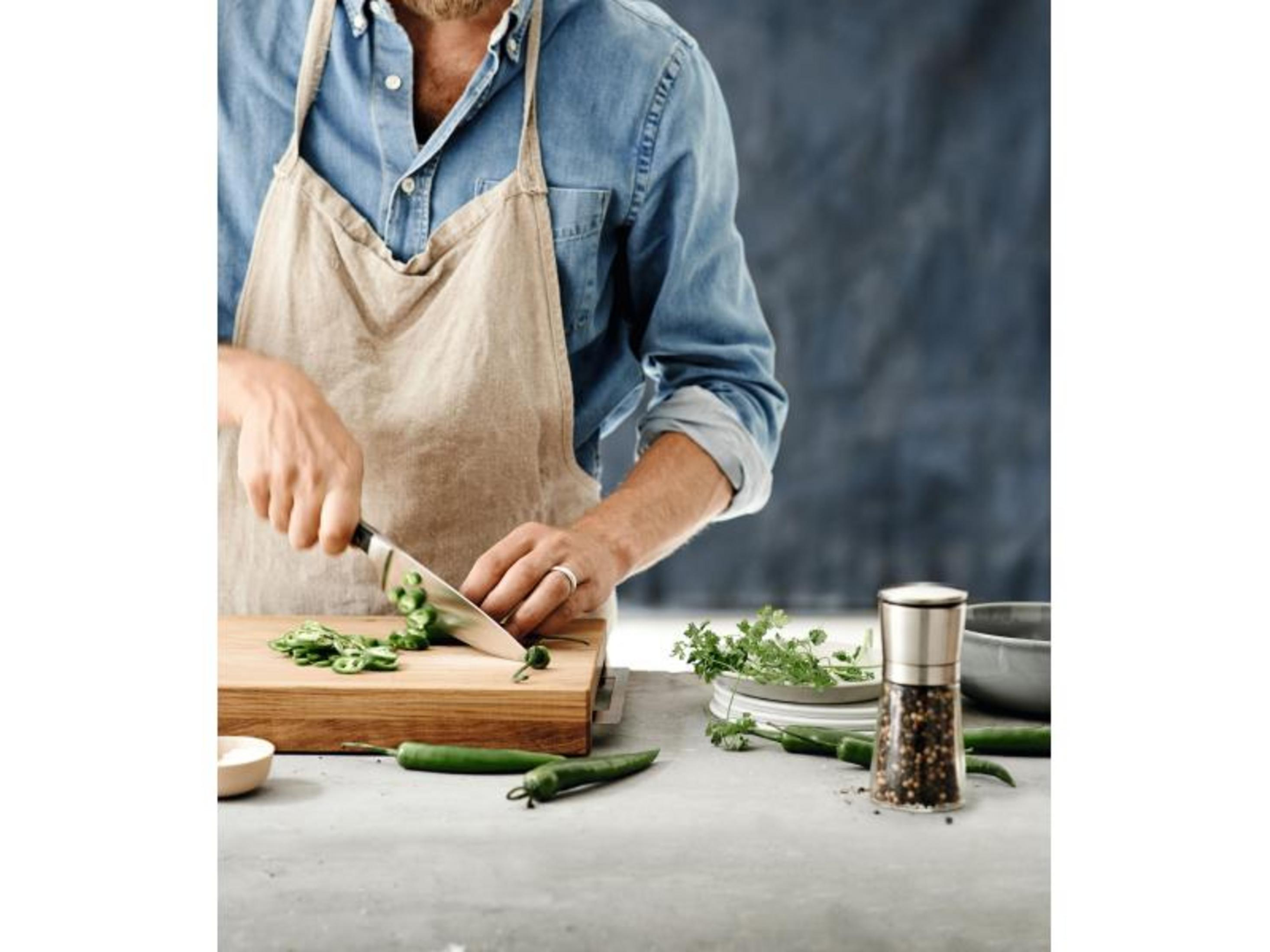 Mężczyzna w dżinsowej koszuli i fartuchu kroi papryczki chili na desce.