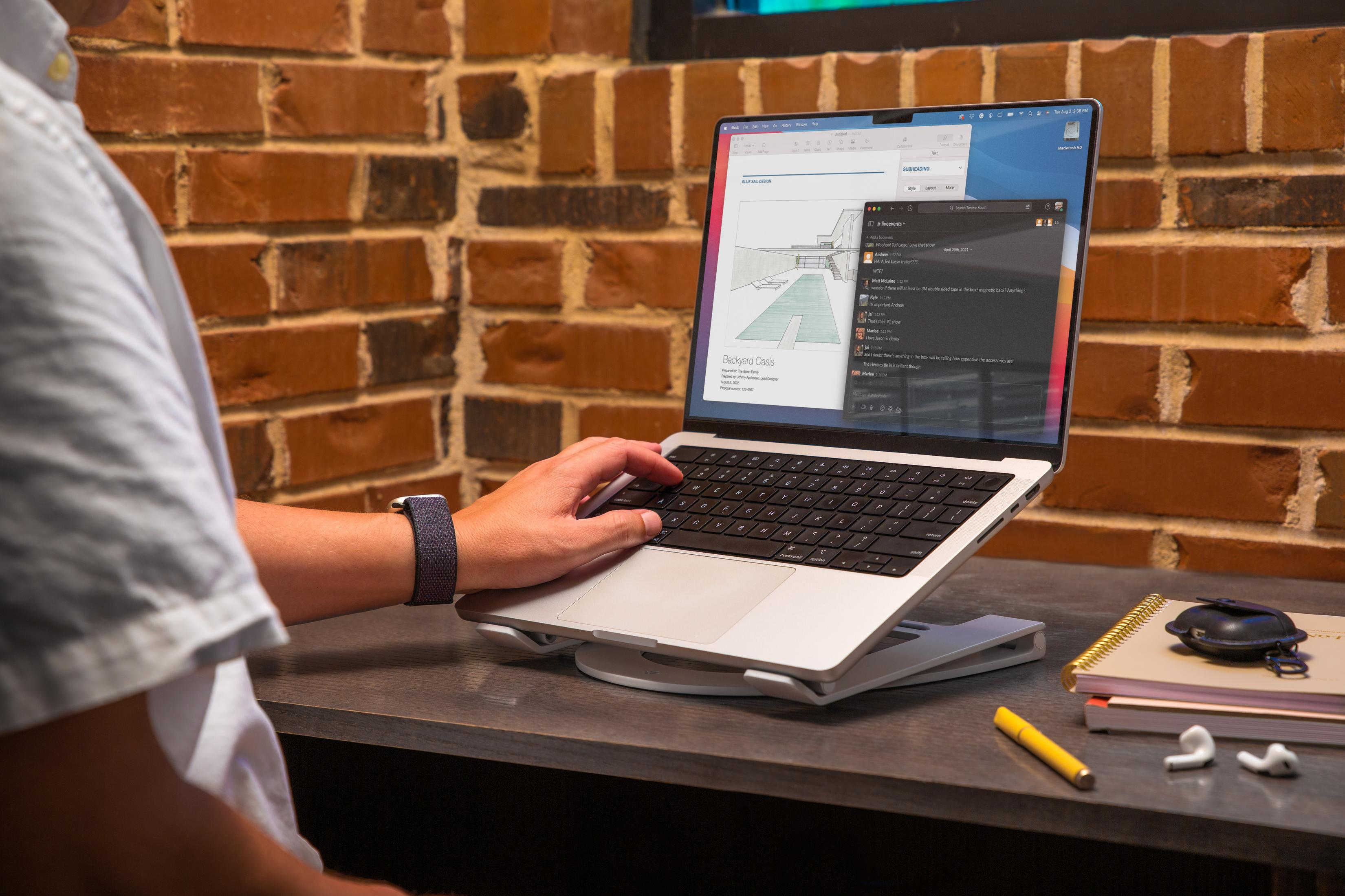 A person types on a laptop on a stand. Brick wall background. Notebook, pen, earbuds on desk.