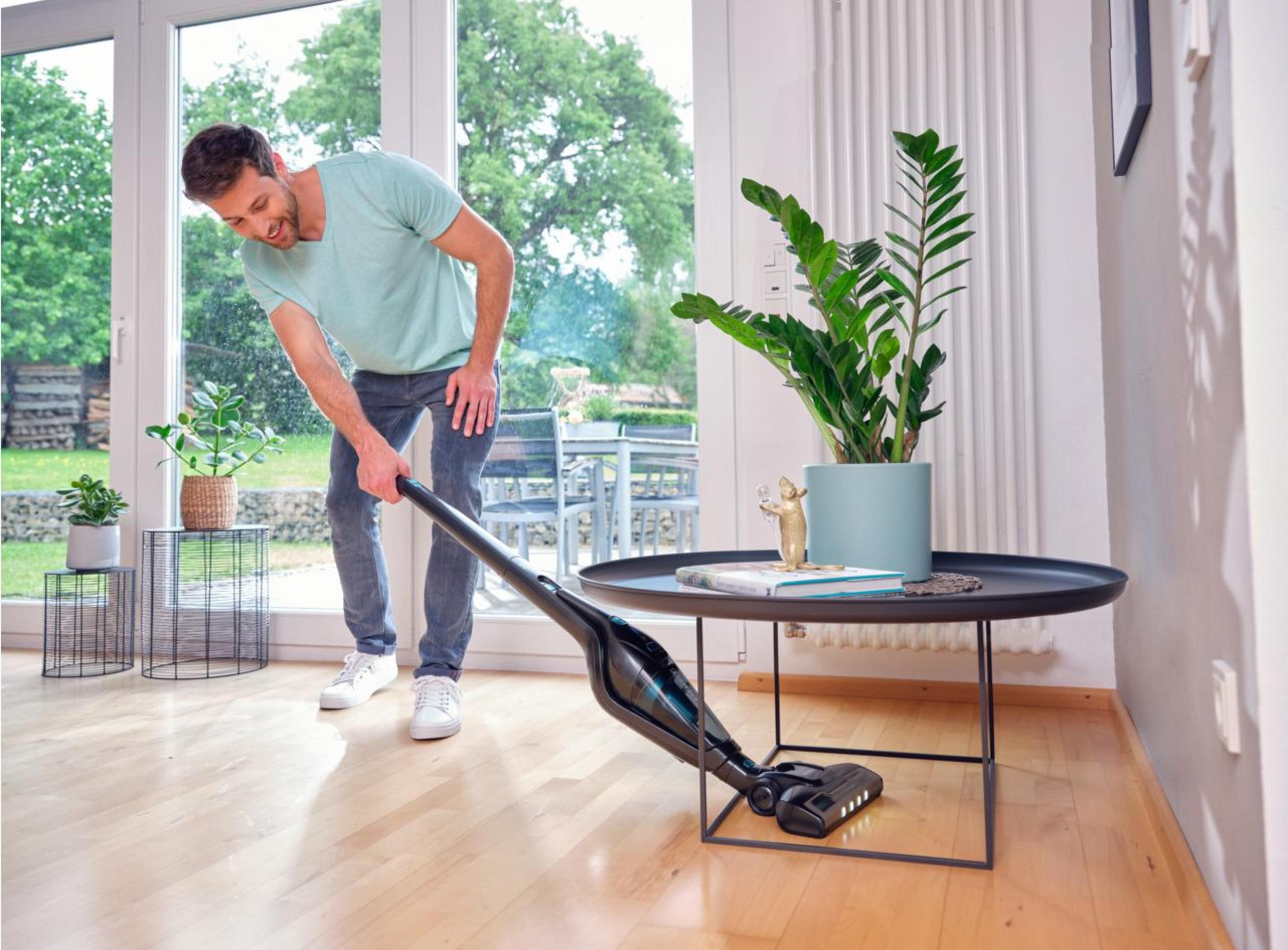 Mężczyzna odkurza drewnianą podłogę czarnym odkurzaczem ręcznym, zielone rośliny z tyłu.