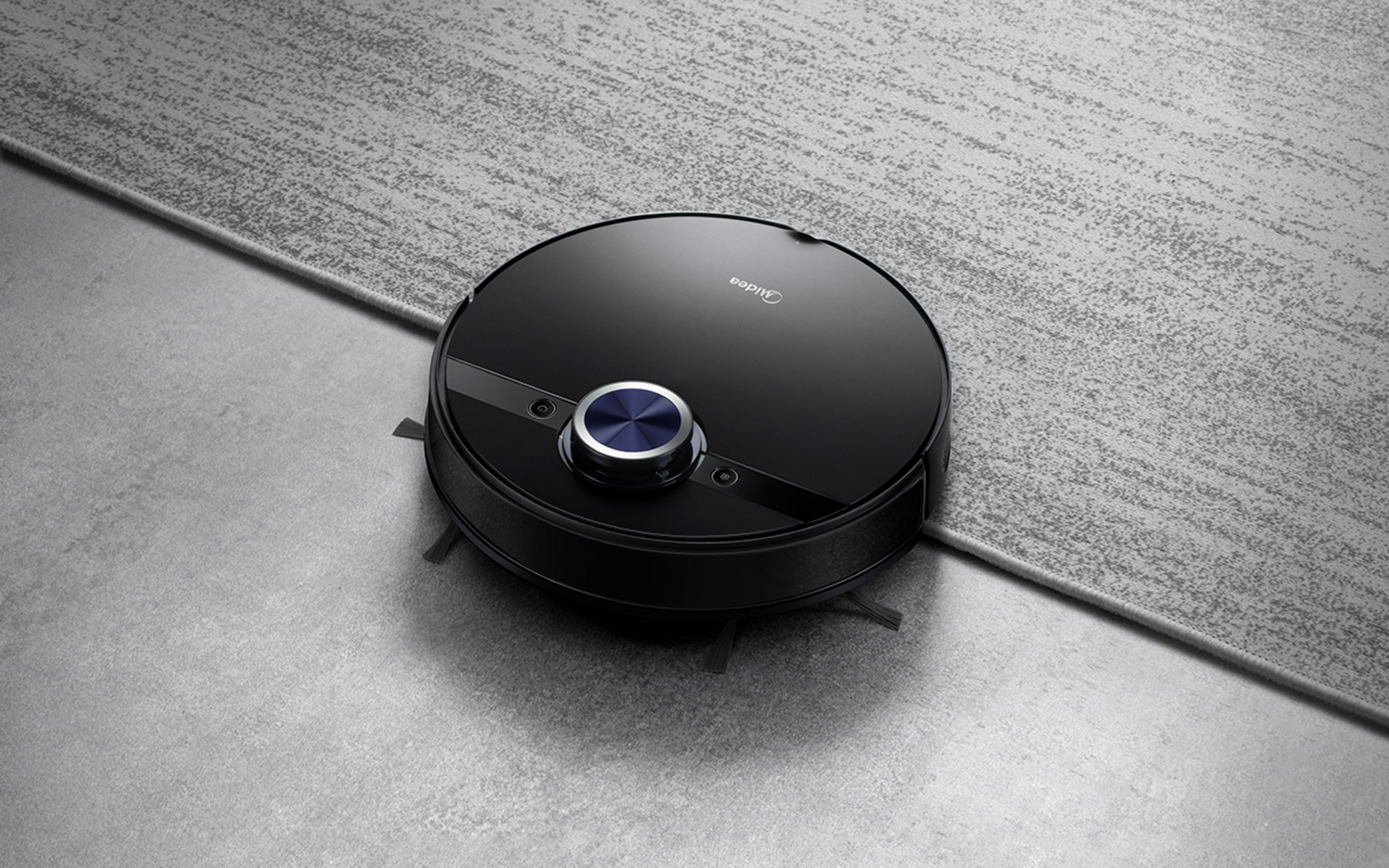 Un robot aspirador negro con botón azul está sobre alfombra gris y suelo de hormigón.