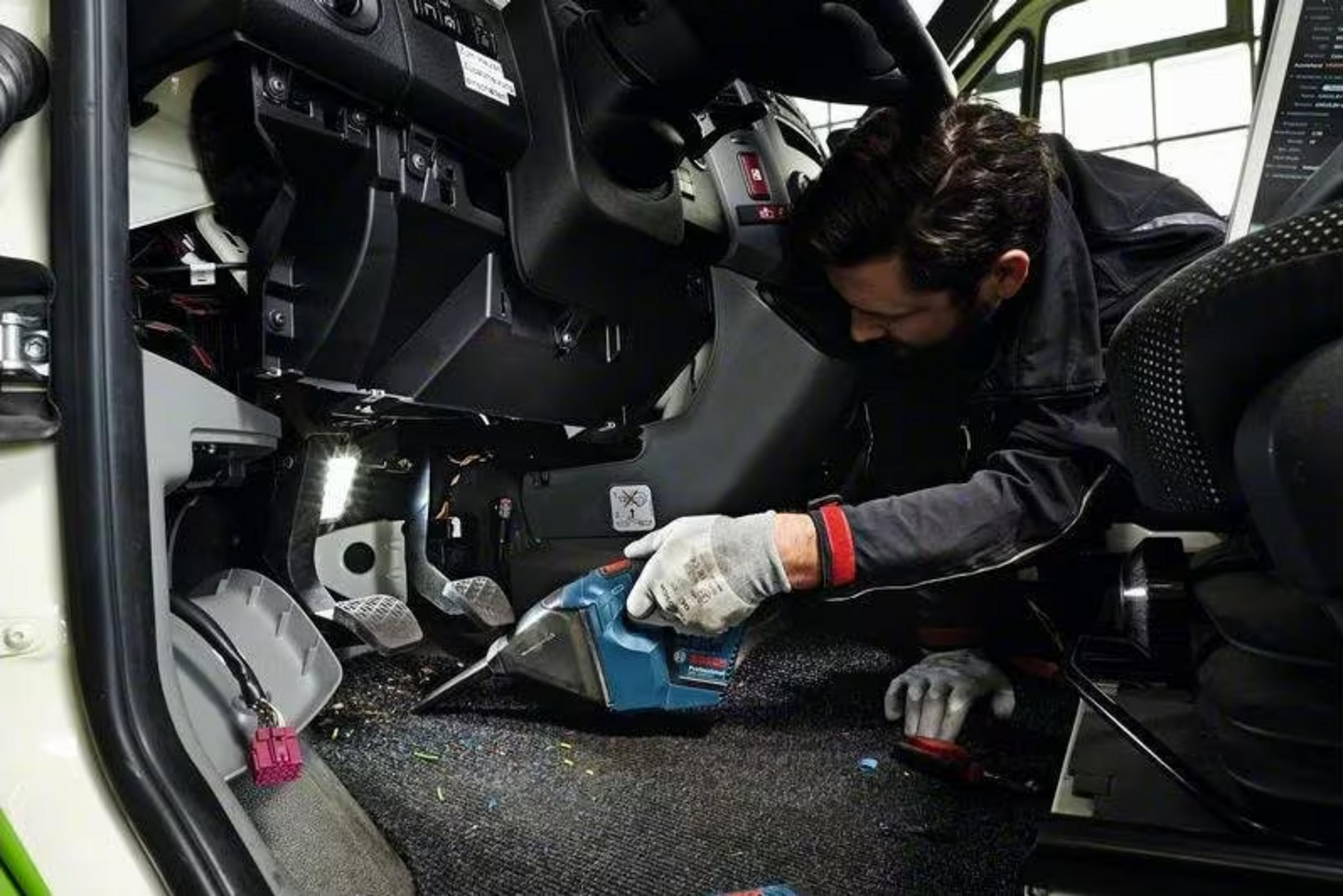 Hombre con guantes limpia suelo de vehículo con aspiradora azul.