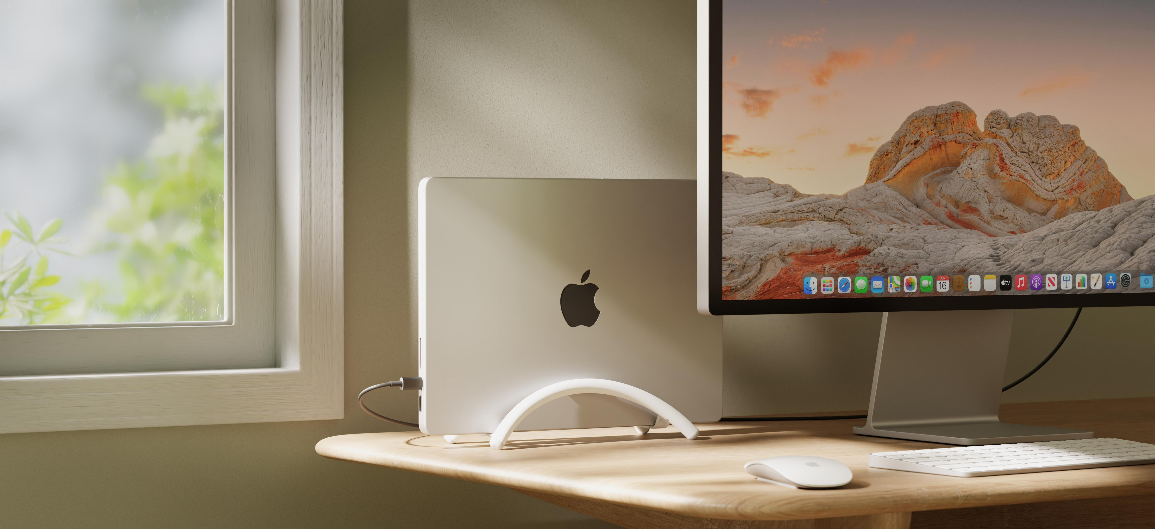 Un laptop Apple argentato su un supporto bianco accanto a un monitor con paesaggio montano.