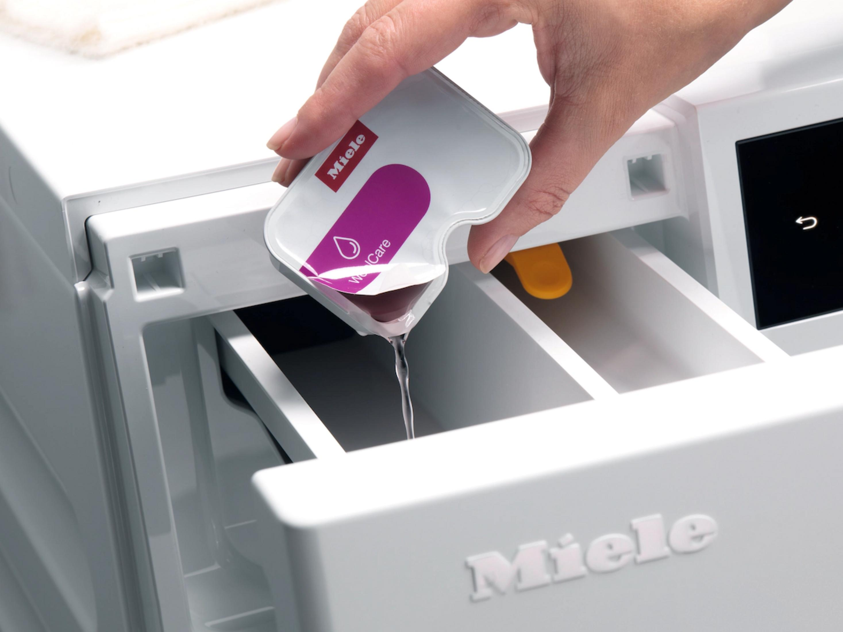 Eine Hand gießt eine Flüssigkeit aus einer Miele-Packung in eine Waschmaschine.