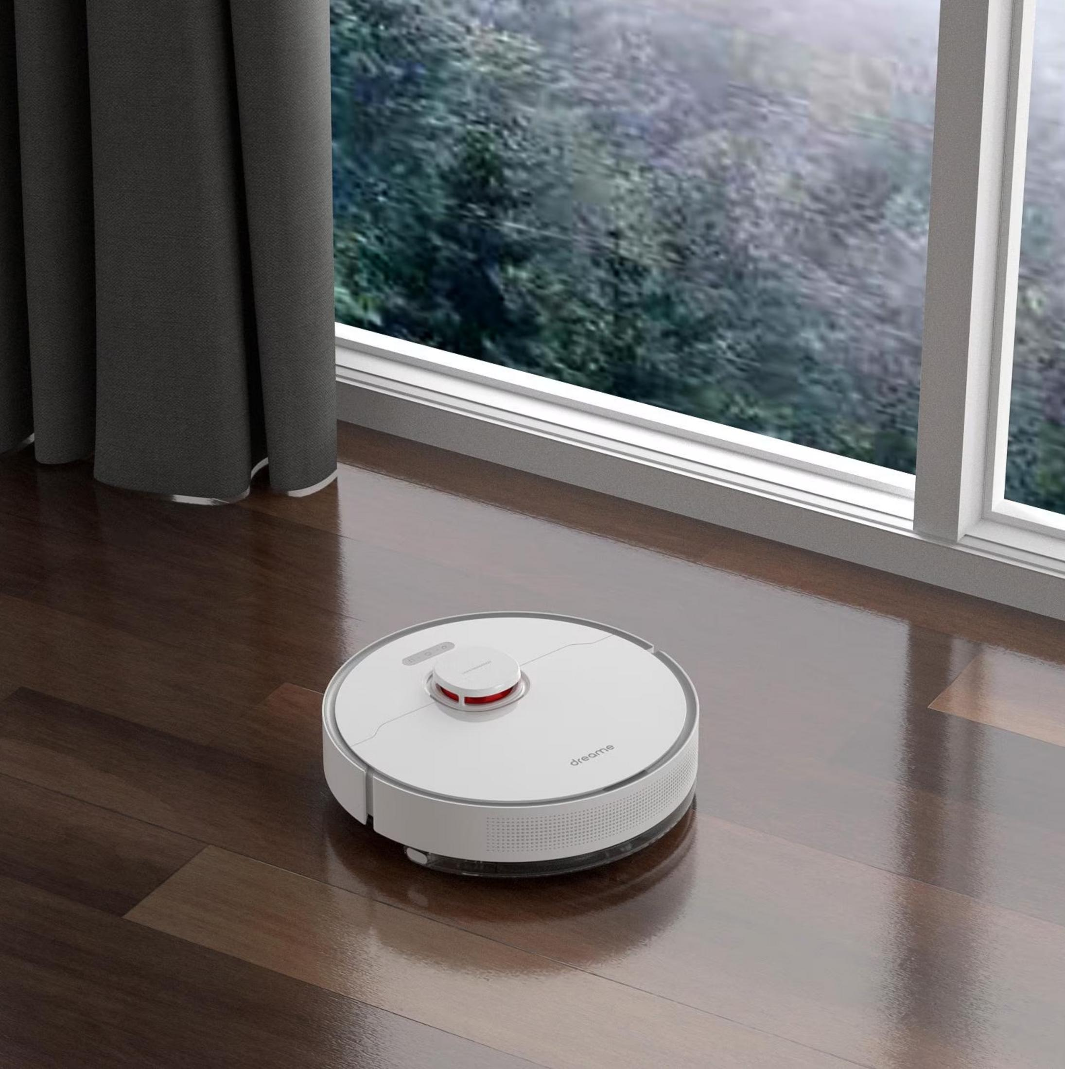 Un aspirateur robot blanc sur un parquet sombre devant une fenêtre avec des arbres.