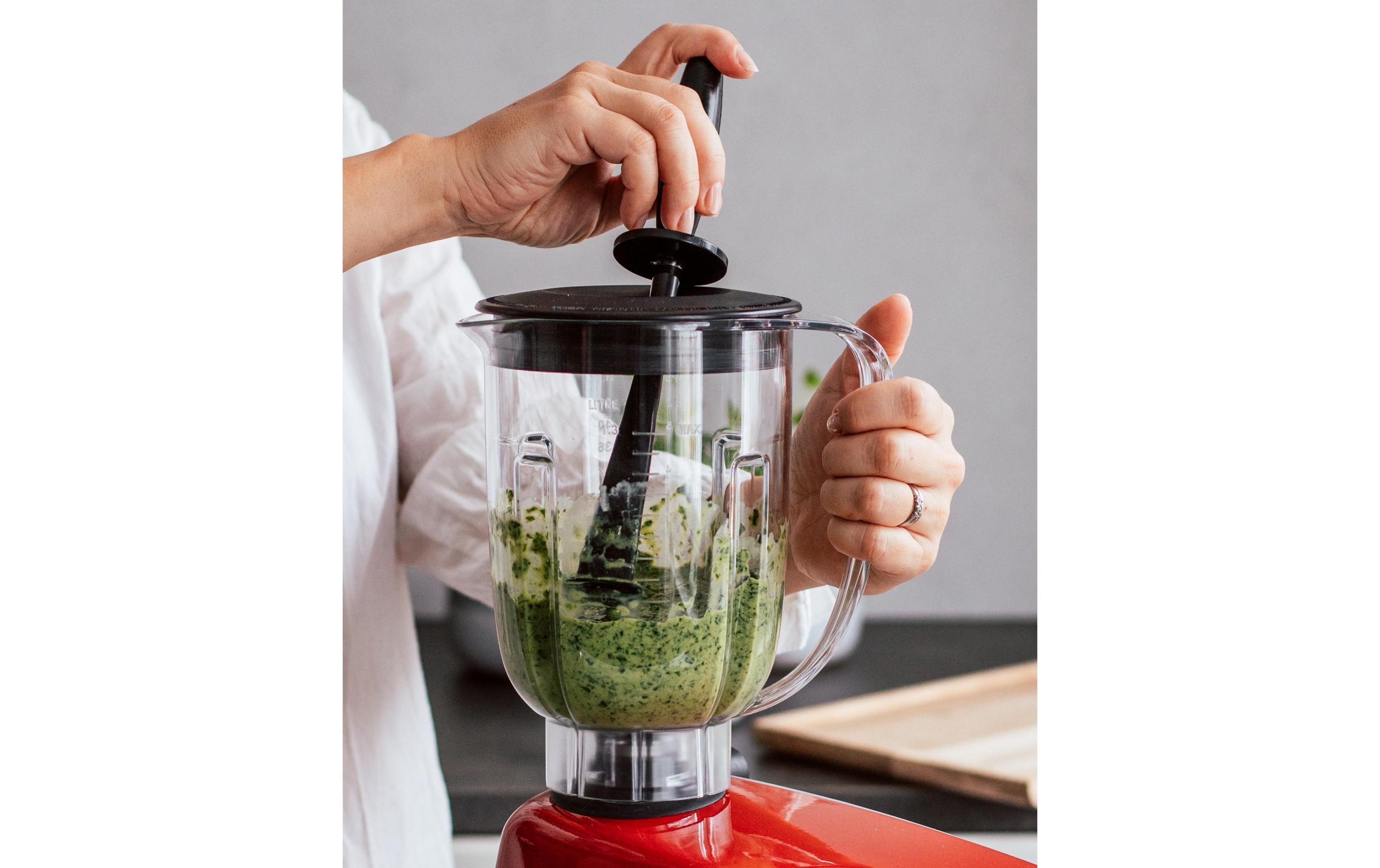 Hände benutzen einen schwarzen Stampfer, um grüne Masse in einem Mixer zu zerdrücken.
