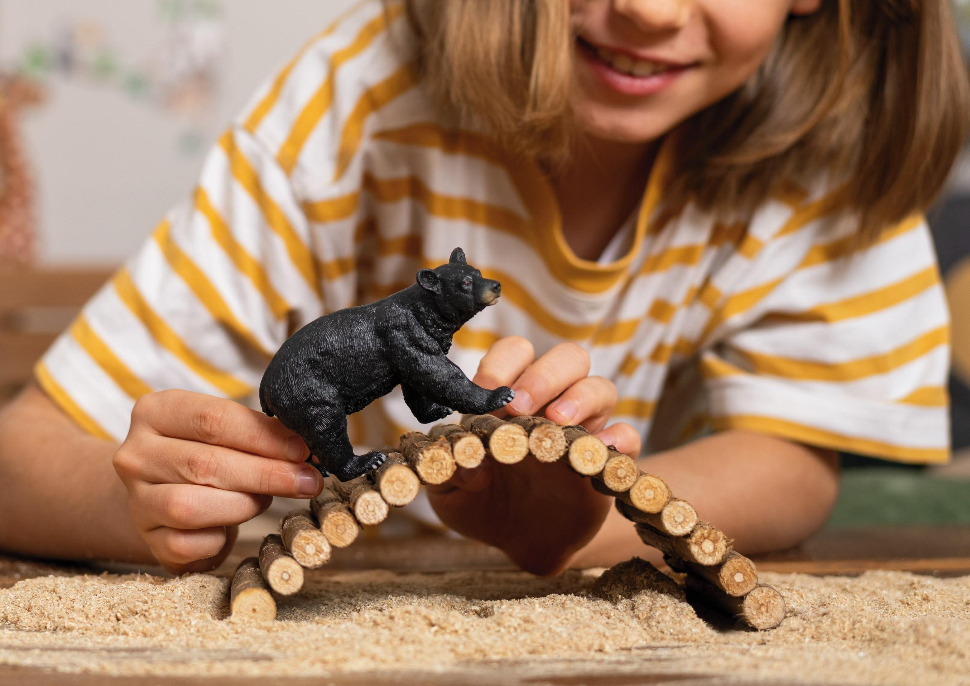 Dziecko w pasiastym T-shircie trzyma figurkę niedźwiedzia na drewnianym moście nad piaskiem.