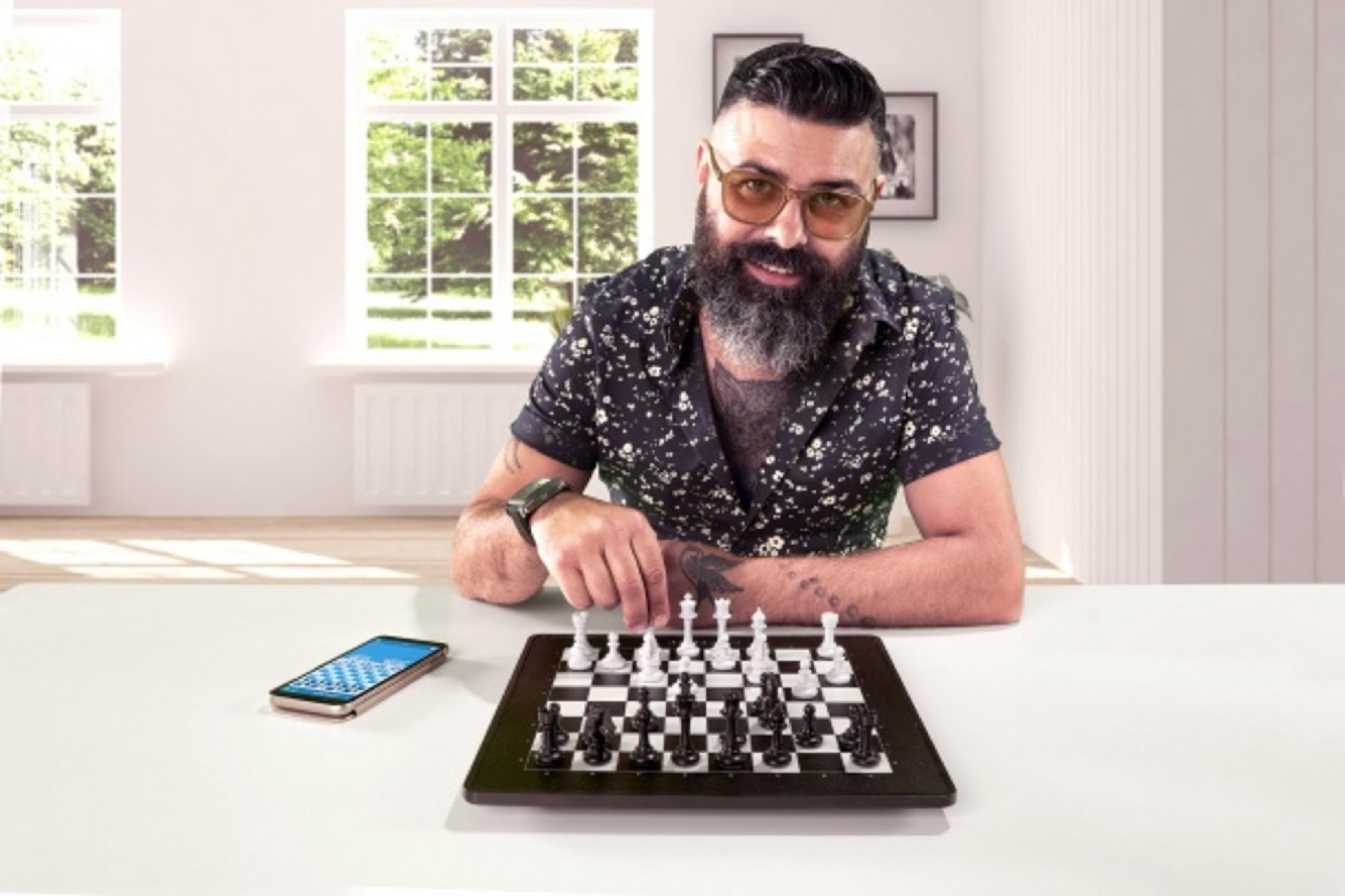 Mann spielt Schach. Ein Handy liegt daneben. Fenster im Hintergrund.