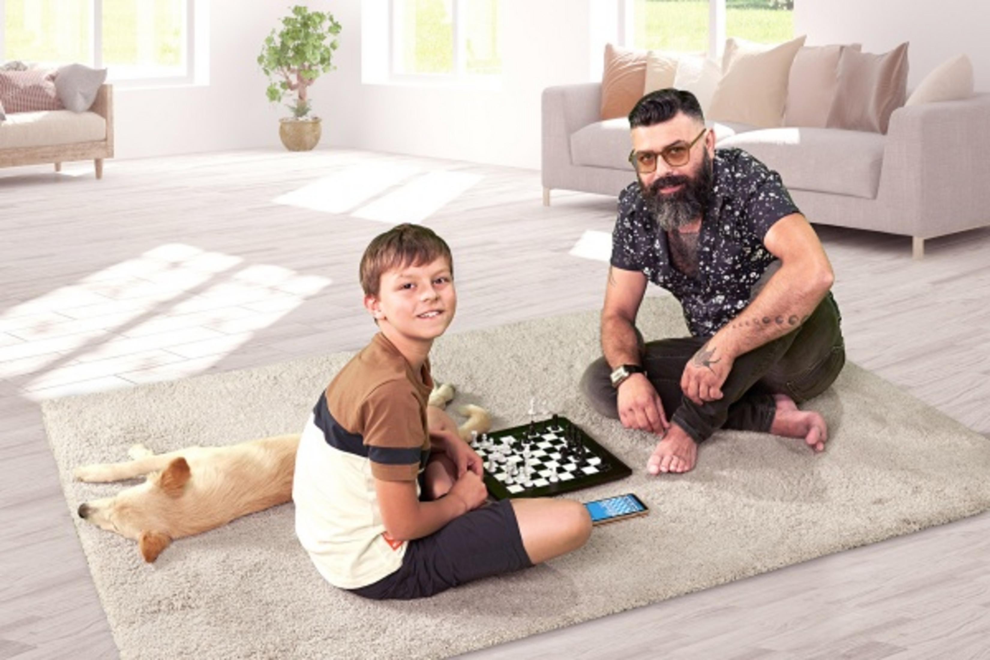 Ein Mann und ein Junge spielen Schach auf einem Teppich. Ein Hund liegt daneben.
