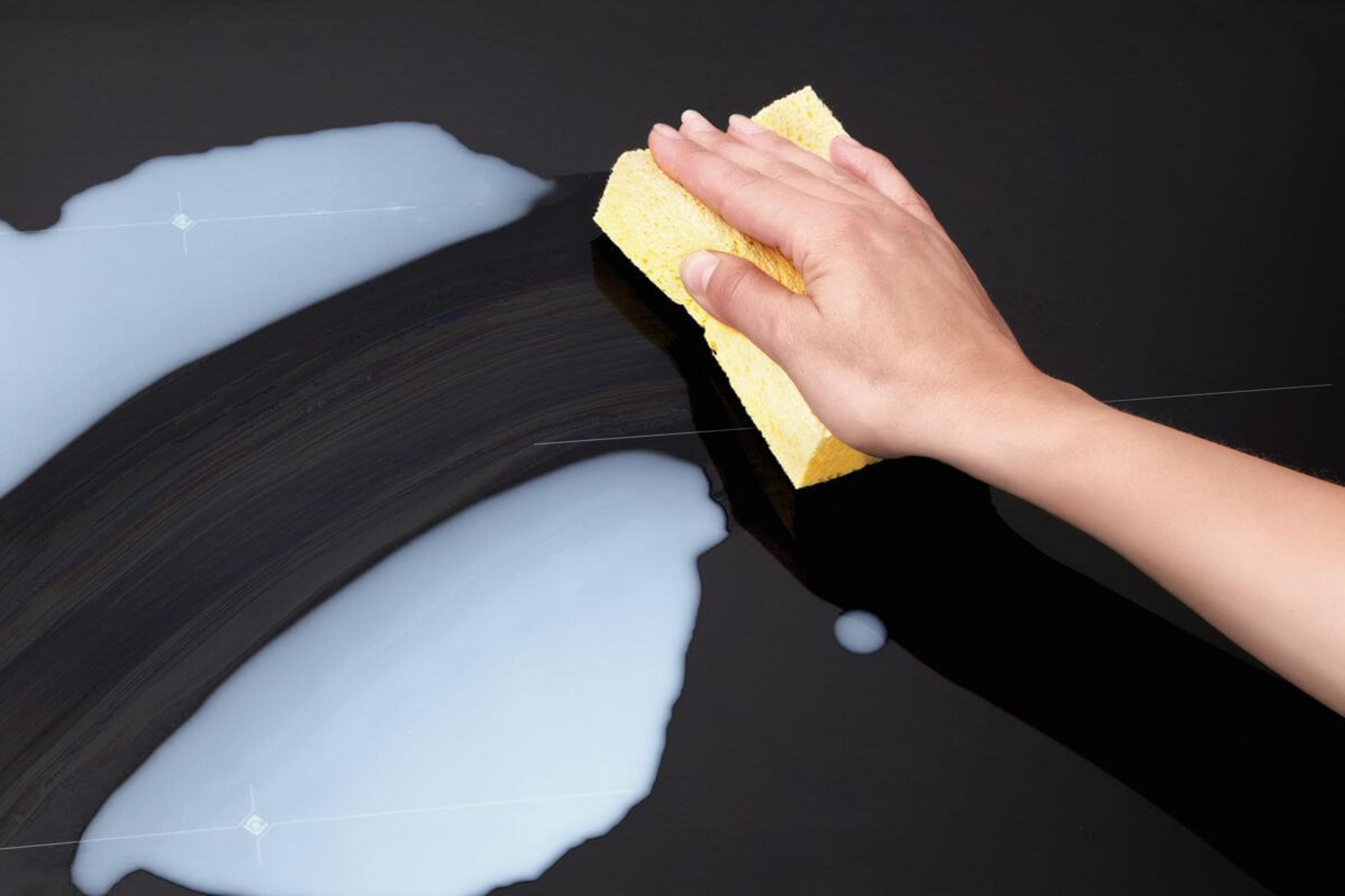 Ręka z żółtą gąbką wyciera czarną powierzchnię z białym płynem.