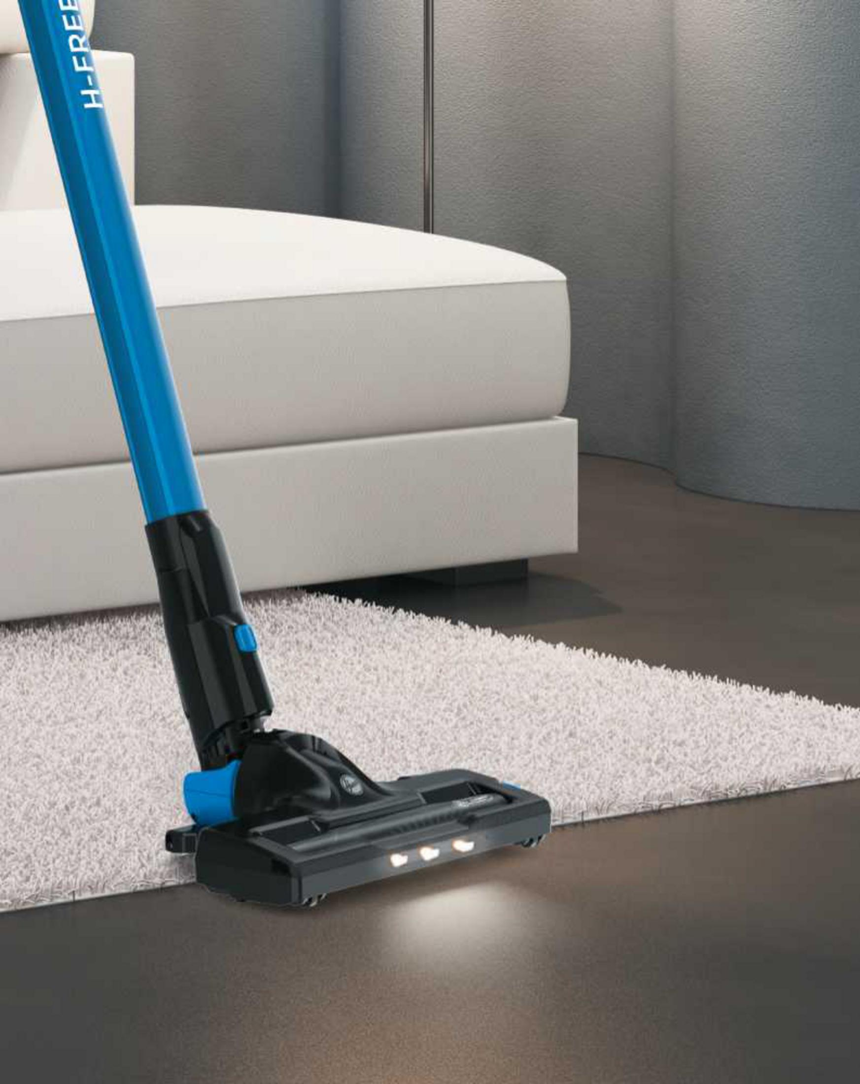 Una aspiradora de palo azul limpia una alfombra clara y suelo oscuro.