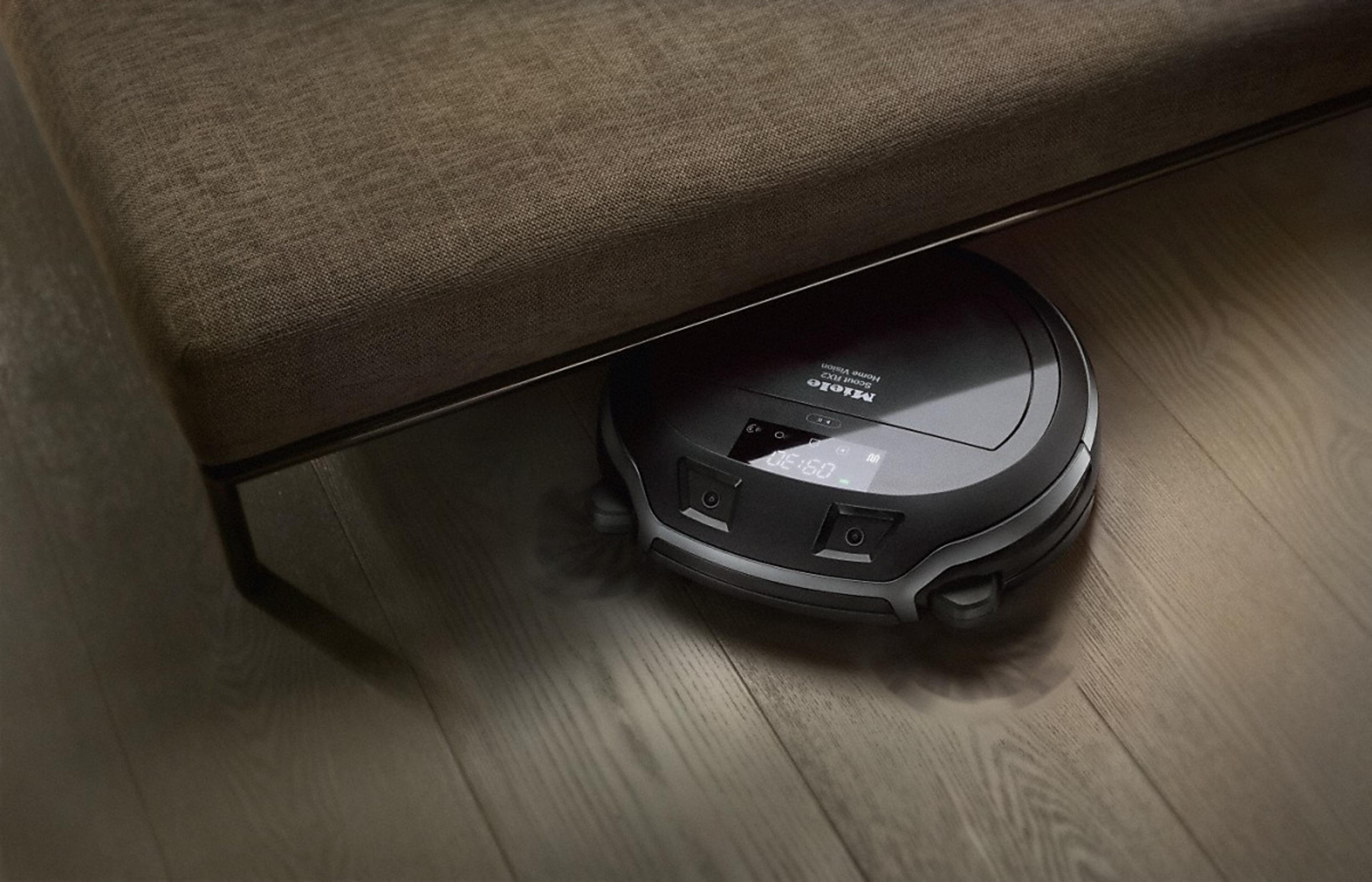 Ein schwarzer Saugroboter unter einem braunen Sofa auf Holzboden.