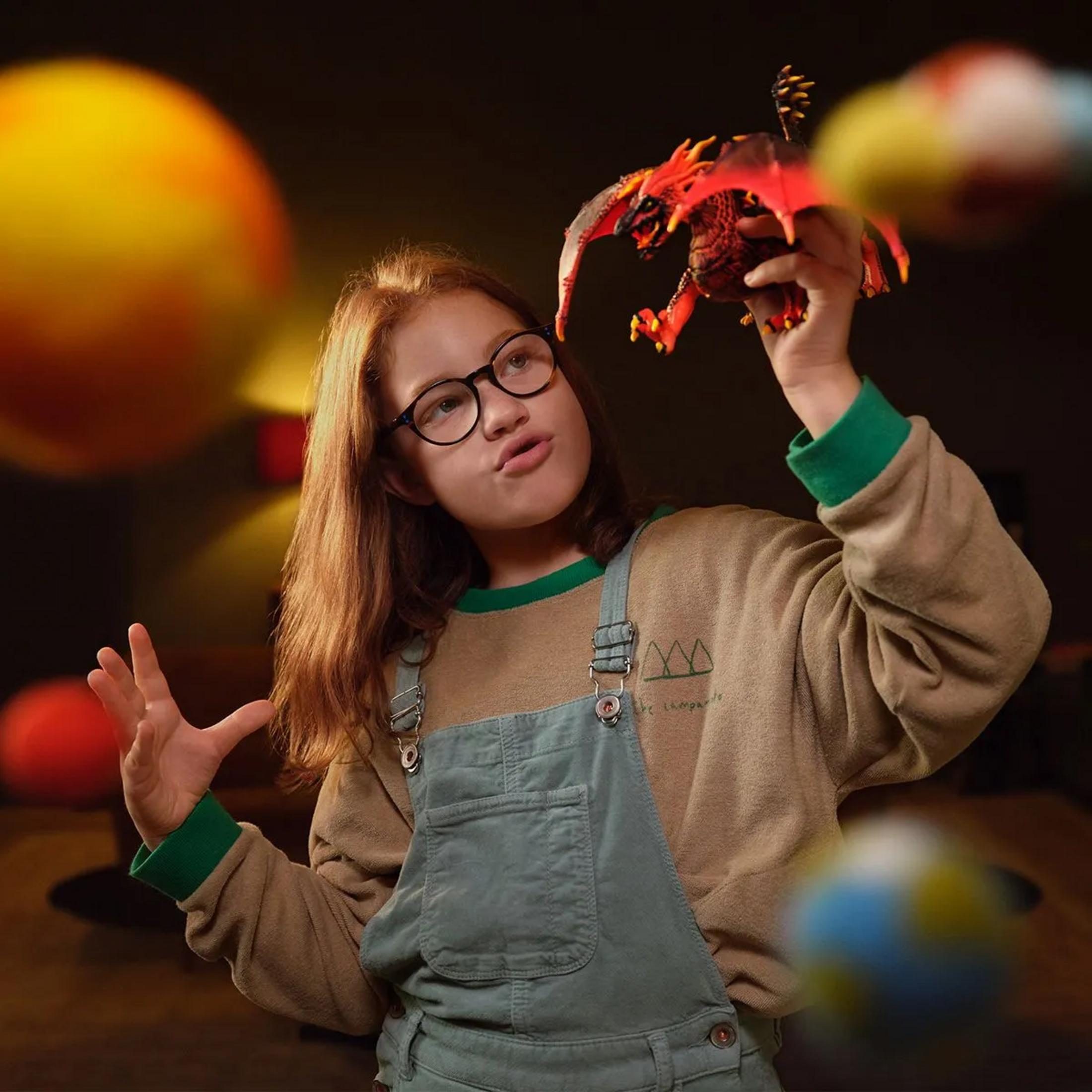 Dziewczynka w okularach trzyma w dłoni czerwoną zabawkę smoka.