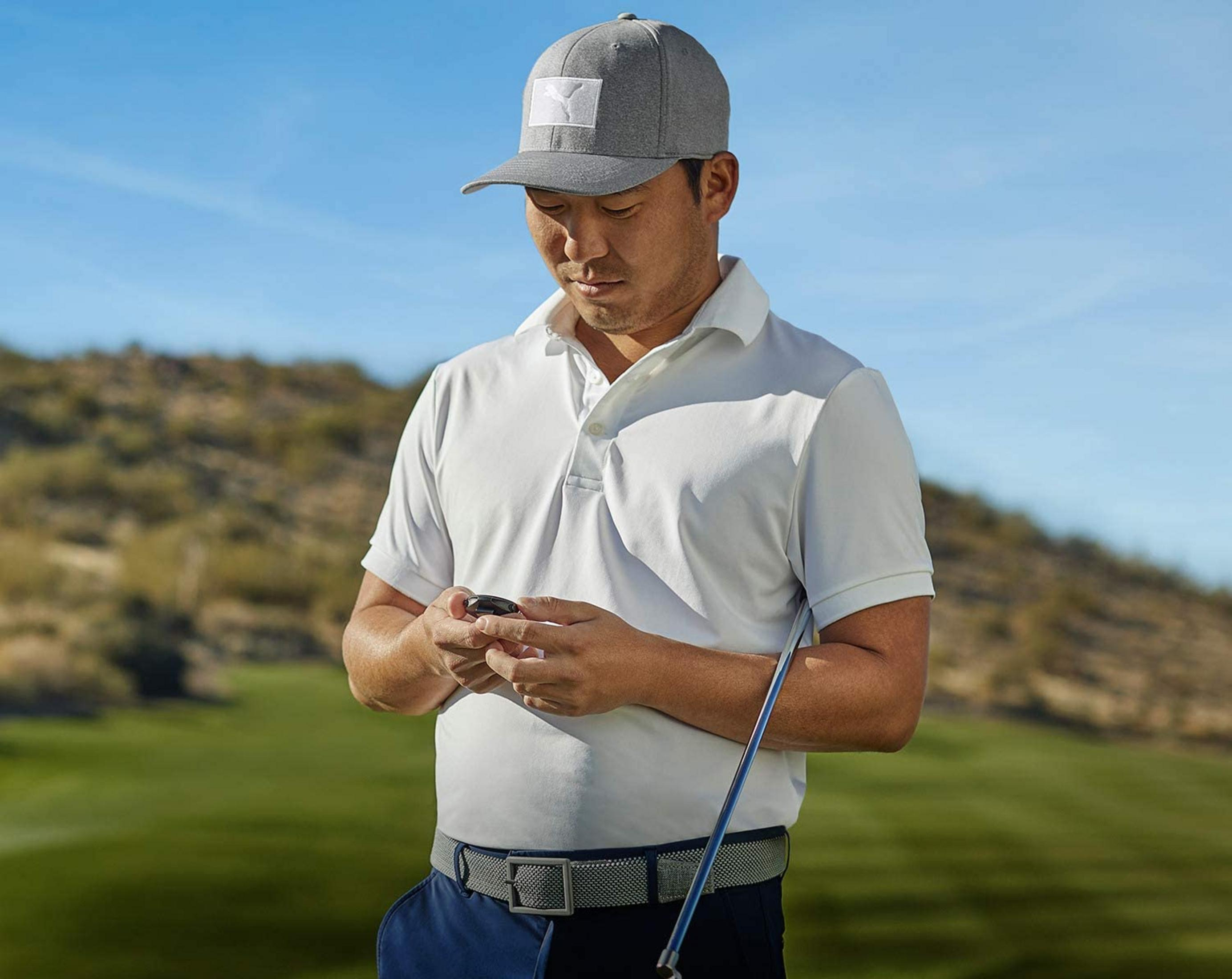 Un golfista con polo blanco y gorra gris revisa su móvil en un campo de golf.