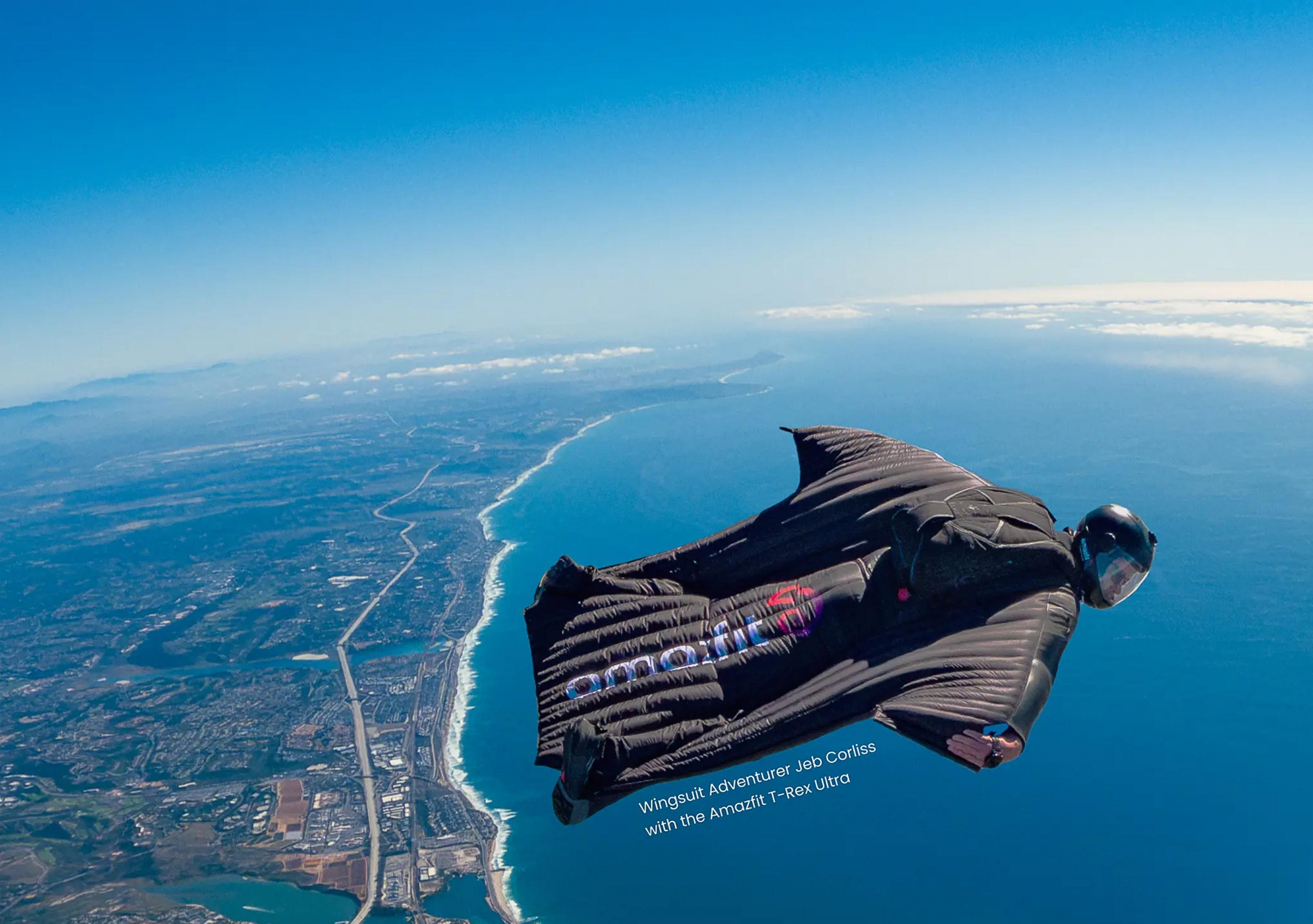 Ein Mann im schwarzen Wingsuit fliegt über der Küste, mit dem Meer rechts.