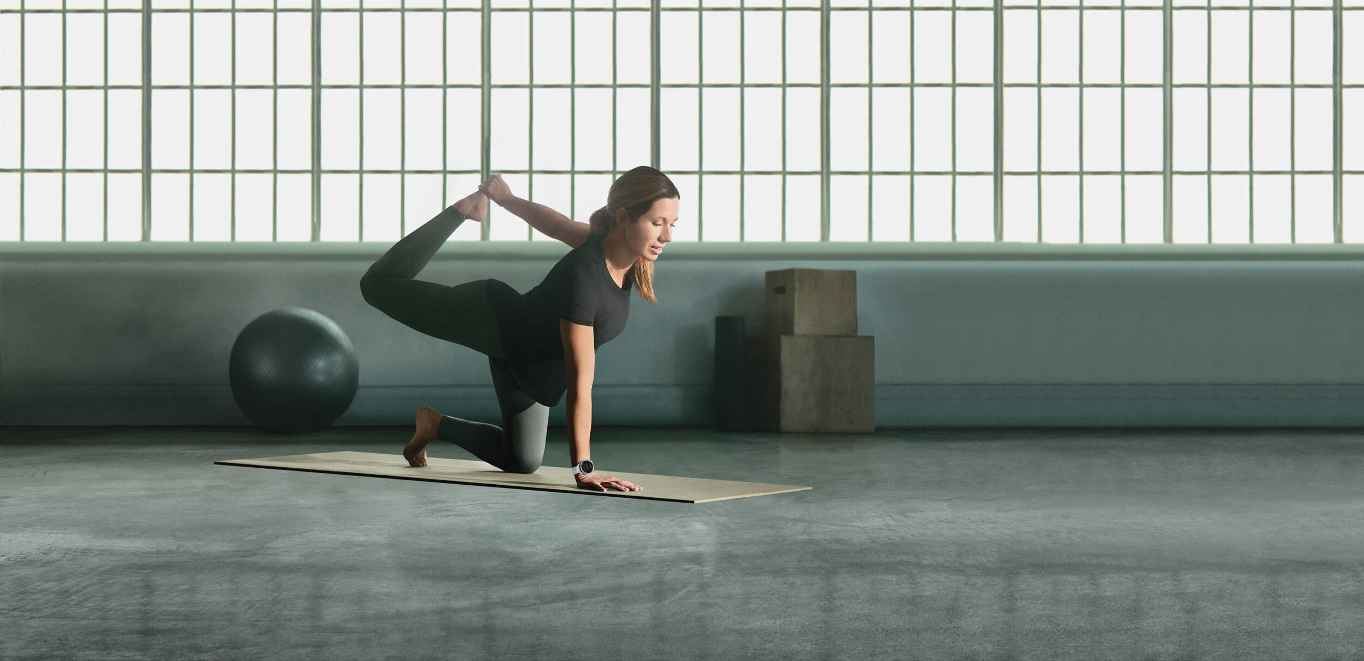 Frau in schwarzer Kleidung macht Yoga auf einer Matte vor einem Fenster.