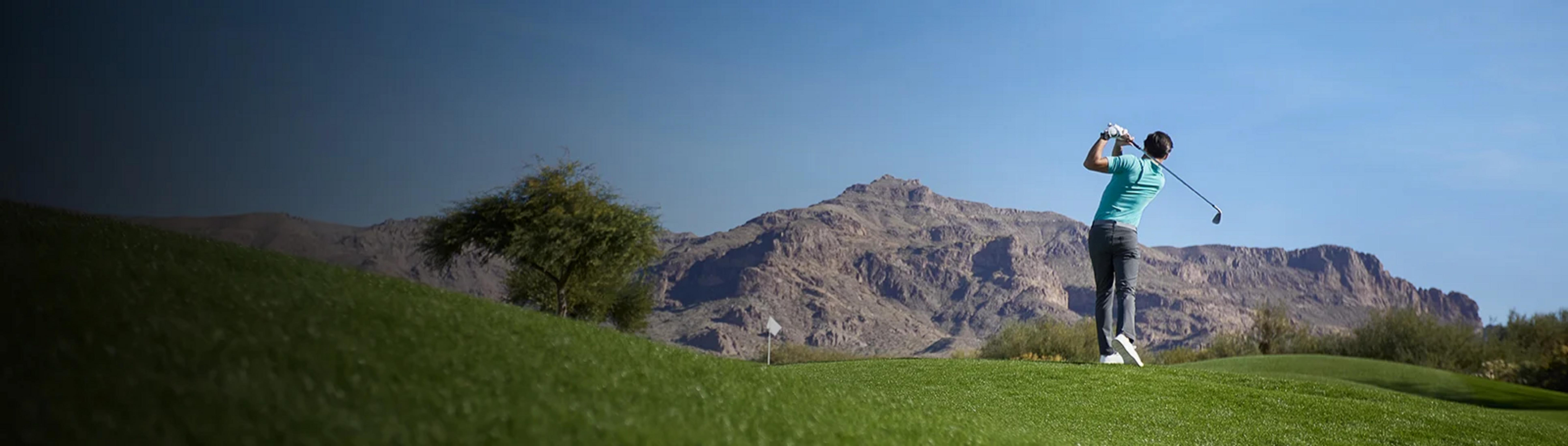 Golfista zamachuje się kijem na bujnym, zielonym polu golfowym z górami w tle.