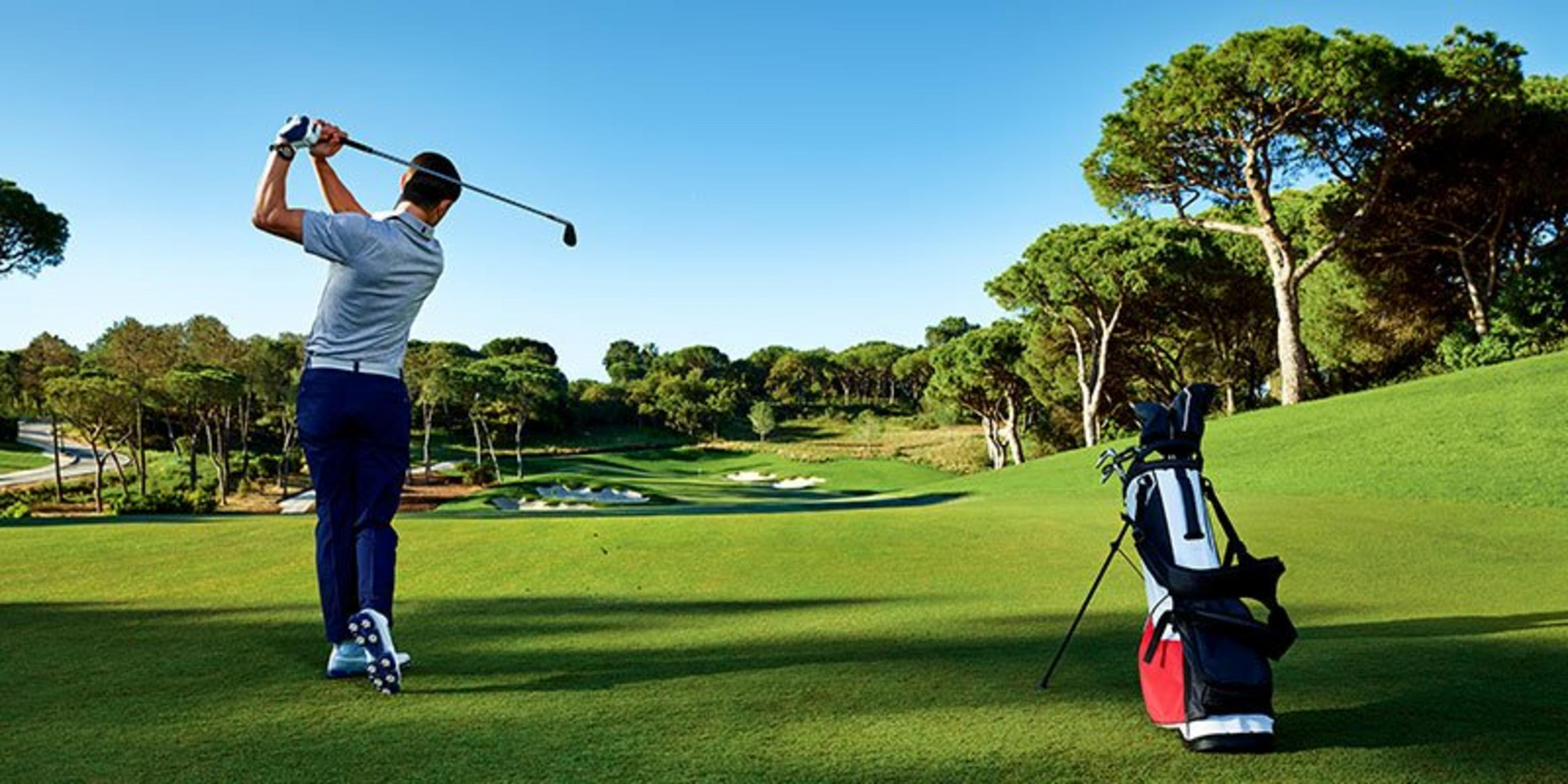 Golfista uderza kijem na zielonym polu golfowym z drzewami i bunkrami.