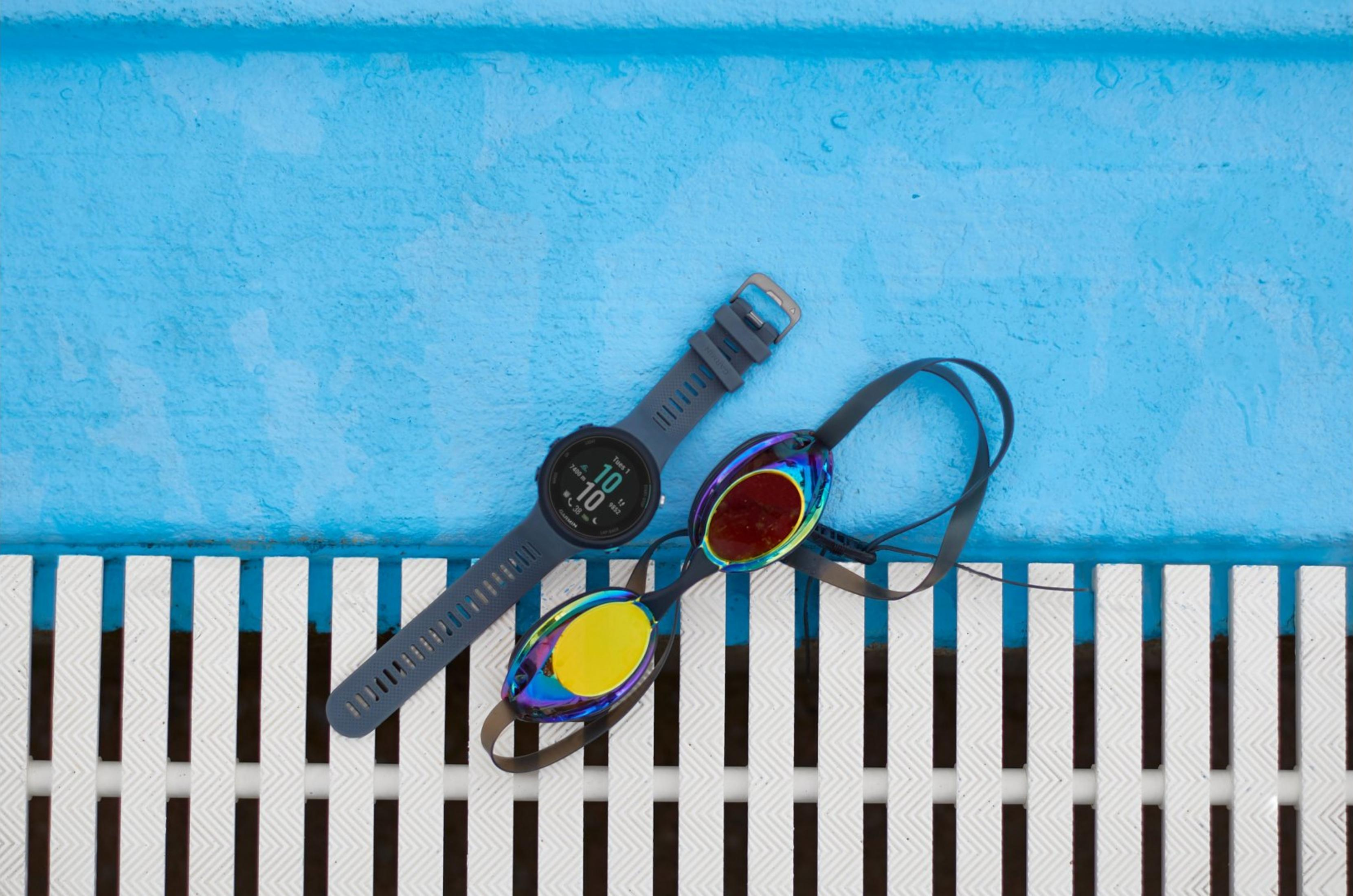 Graue Schwimmuhr und bunte Schwimmbrille liegen auf blauem Wasser und weißem Gitter.