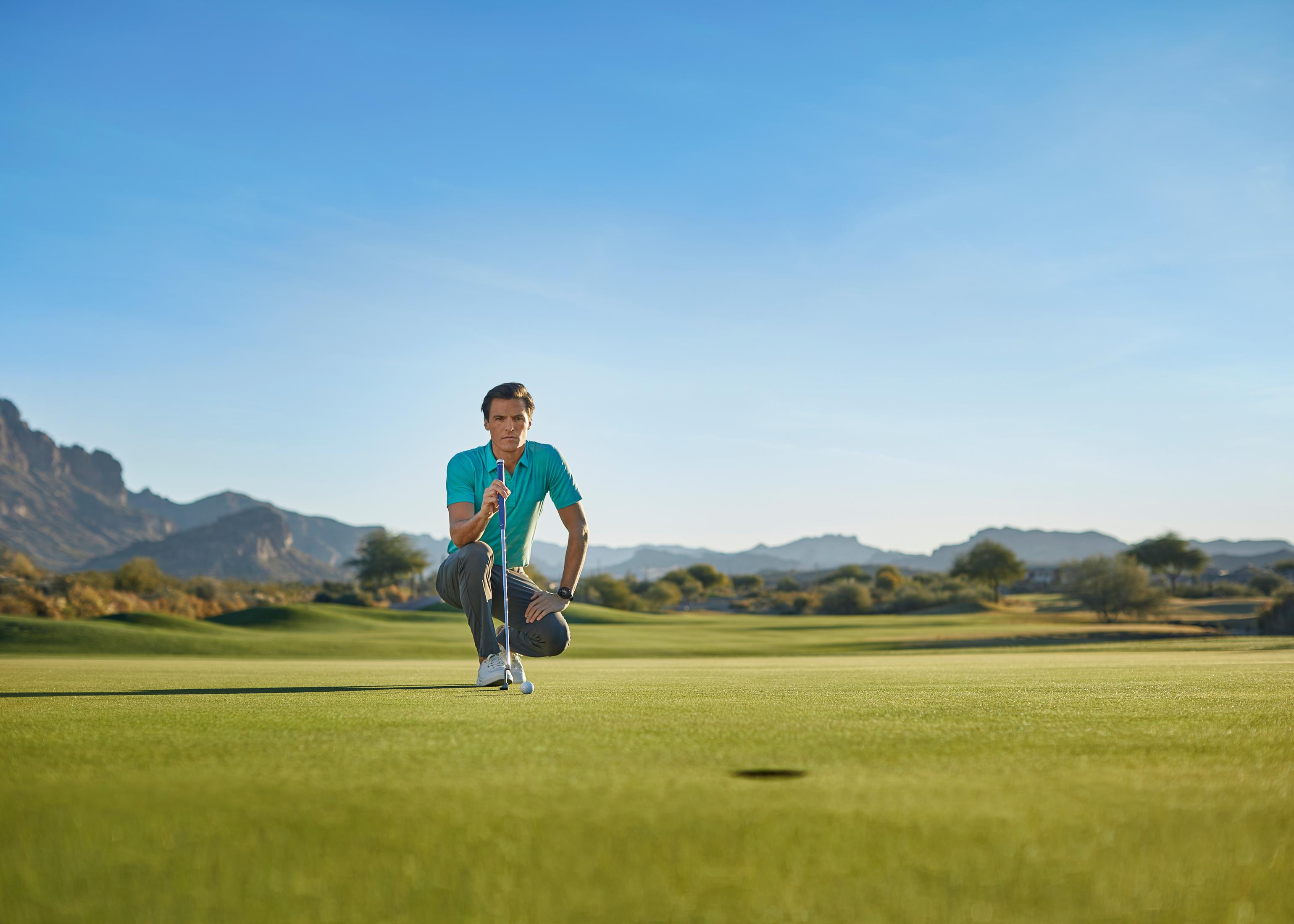 Golfista klęczy na polu, trzymając kij. W tle góry.