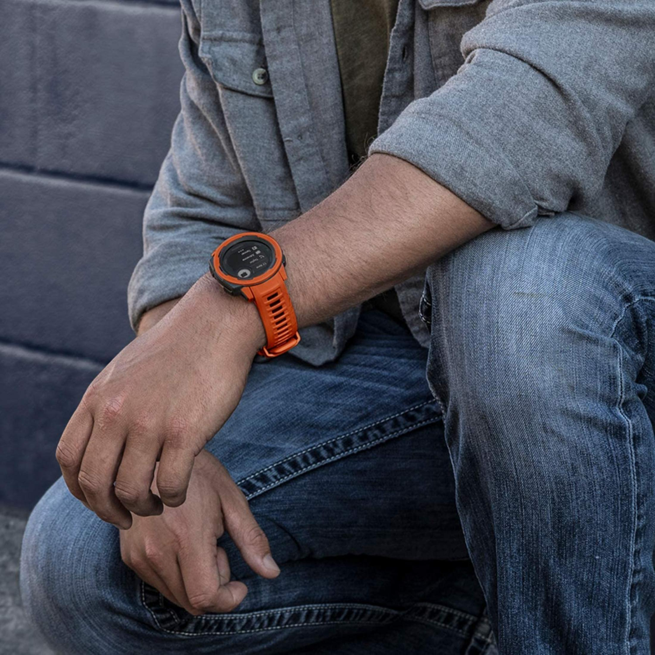 Ein Mann trägt eine orangefarbene Smartwatch am linken Handgelenk, blaue Jeans und eine graue Jacke.