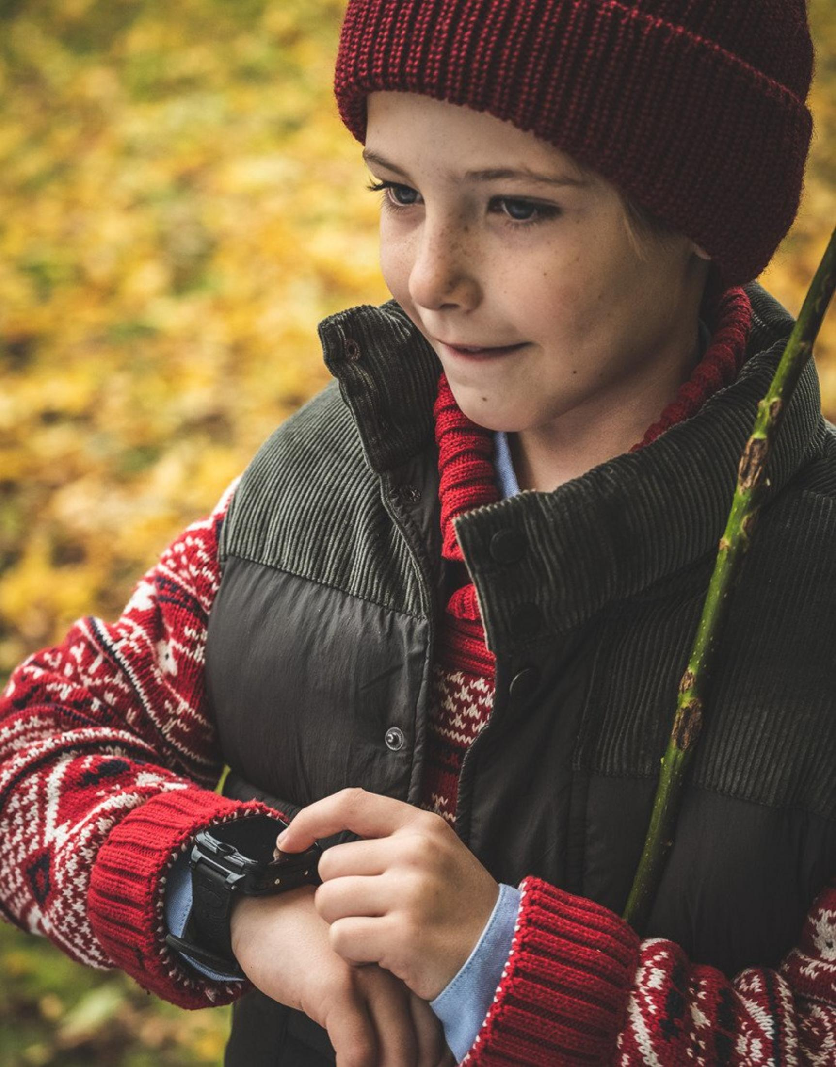 Junge mit roter Mütze und Strickpullover schaut auf seine Uhr.