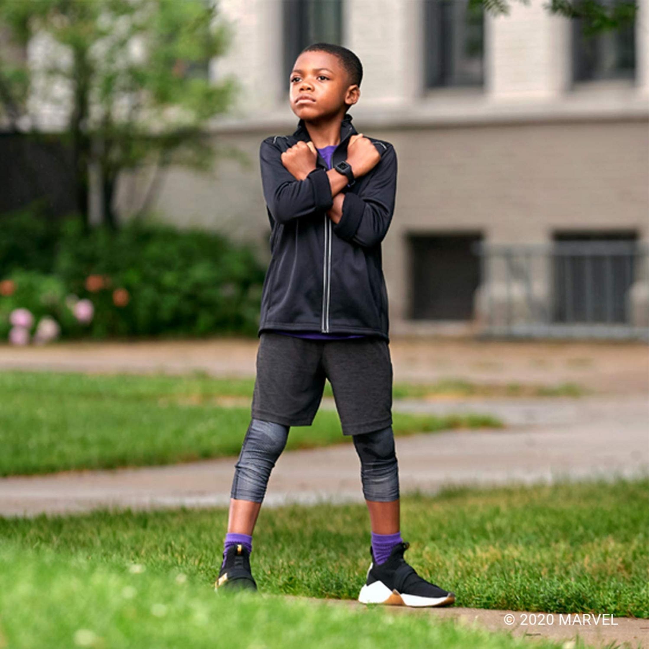 Chłopiec w czarnym stroju stoi na trawie, z rękami skrzyżowanymi na piersi.