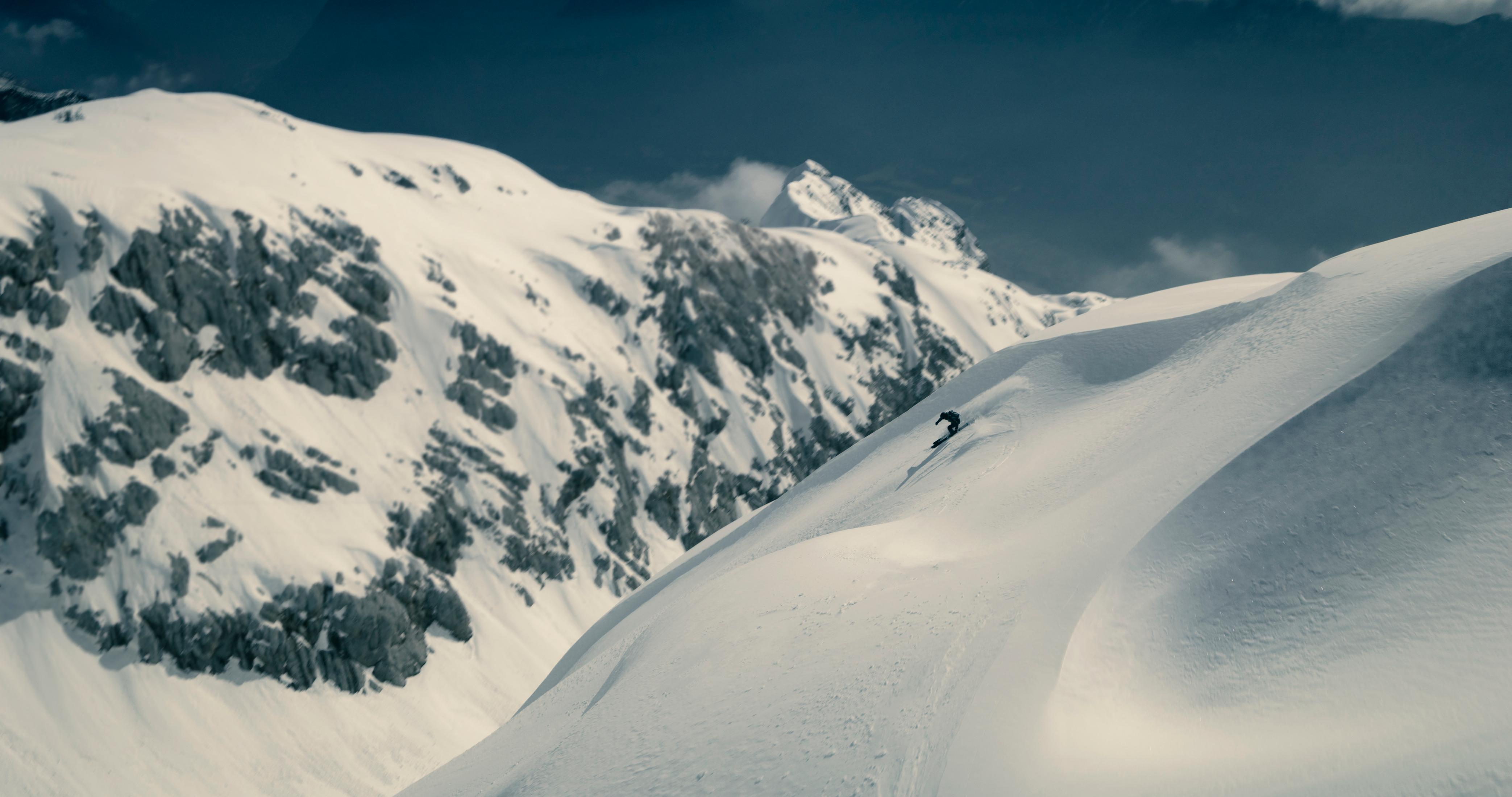 Snowboarder zjeżdża po zaśnieżonym stoku, góry w tle.