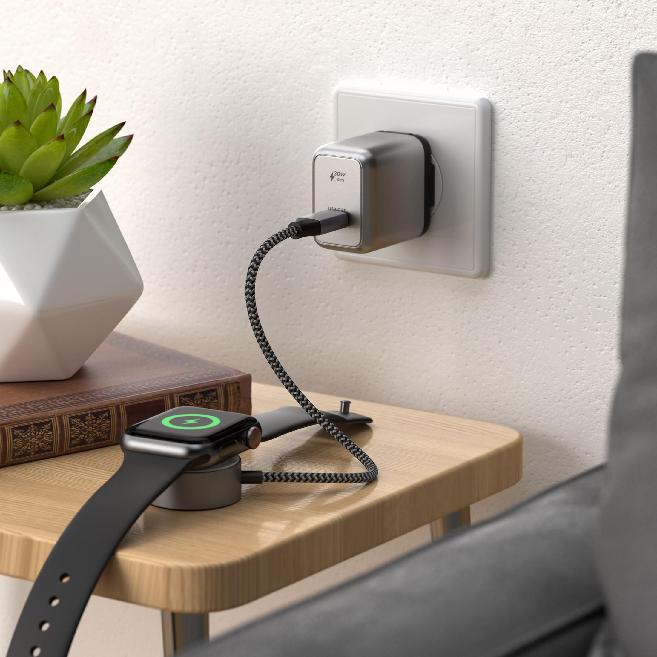 Un caricatore GaN argentato da 30W alimenta un Apple Watch su un comodino.