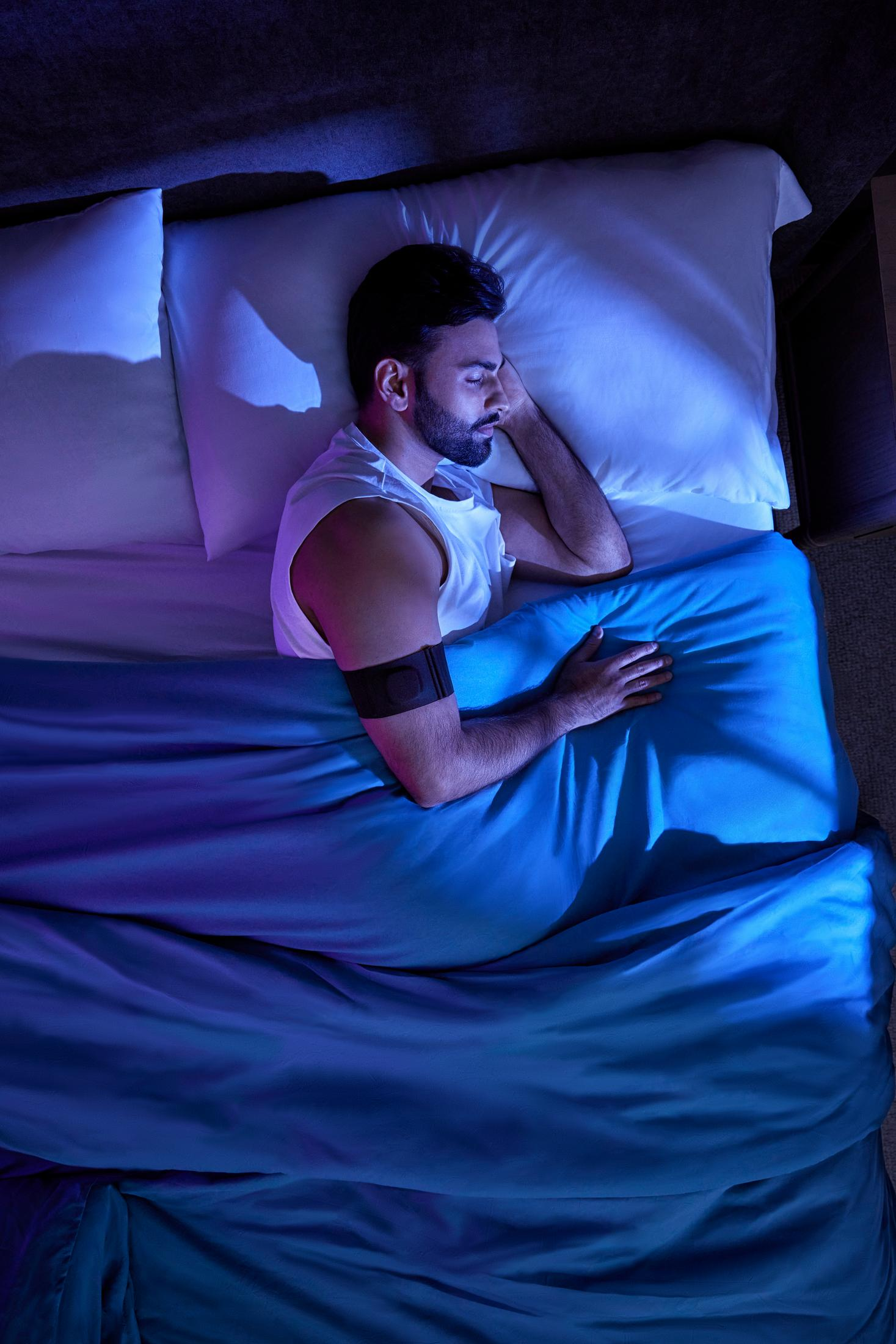 Mann schläft im Bett unter blauer Decke mit Gerät am Arm.