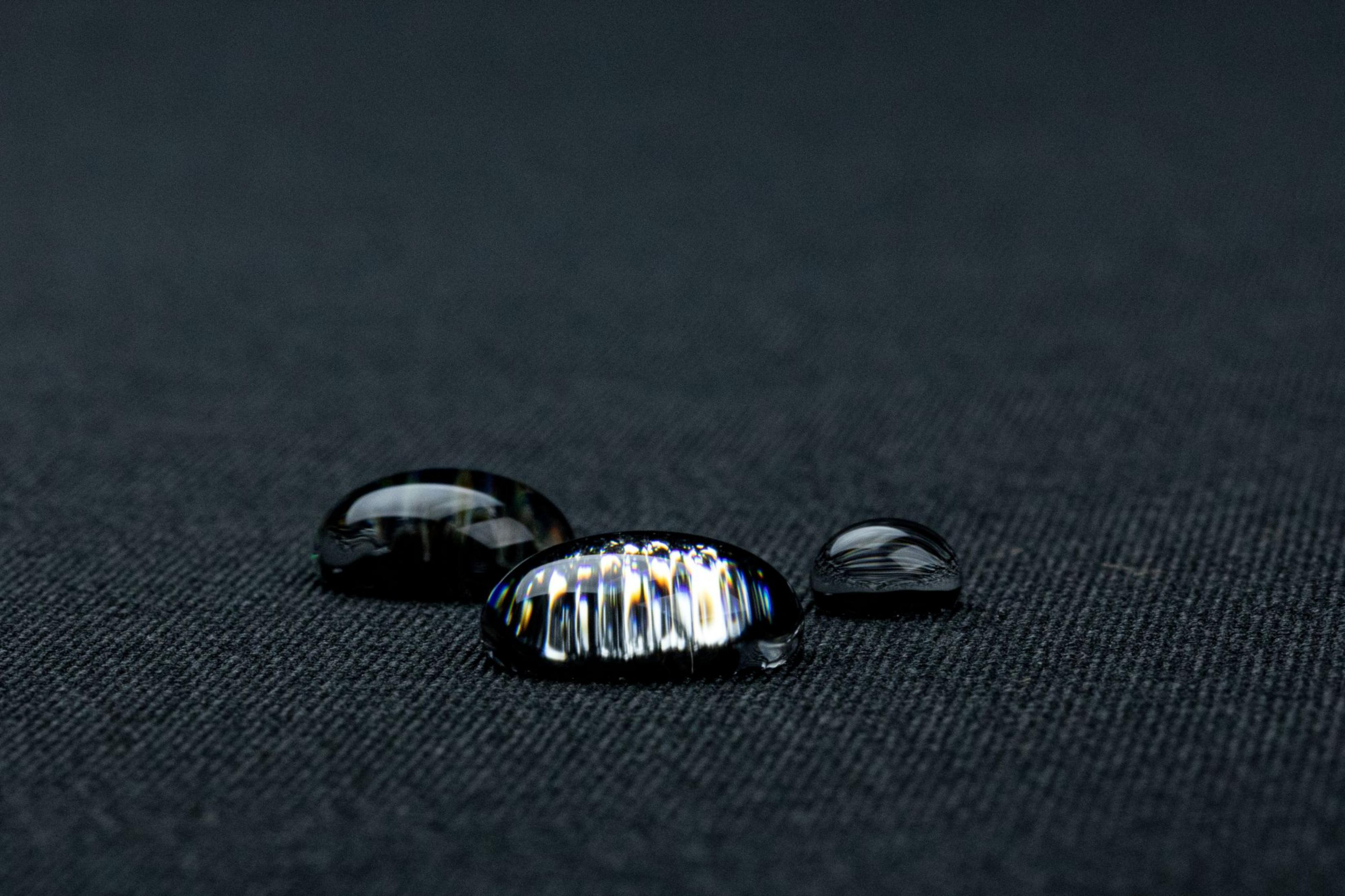 Tre gocce d'acqua lucide su tessuto scuro, con riflessi di luce.