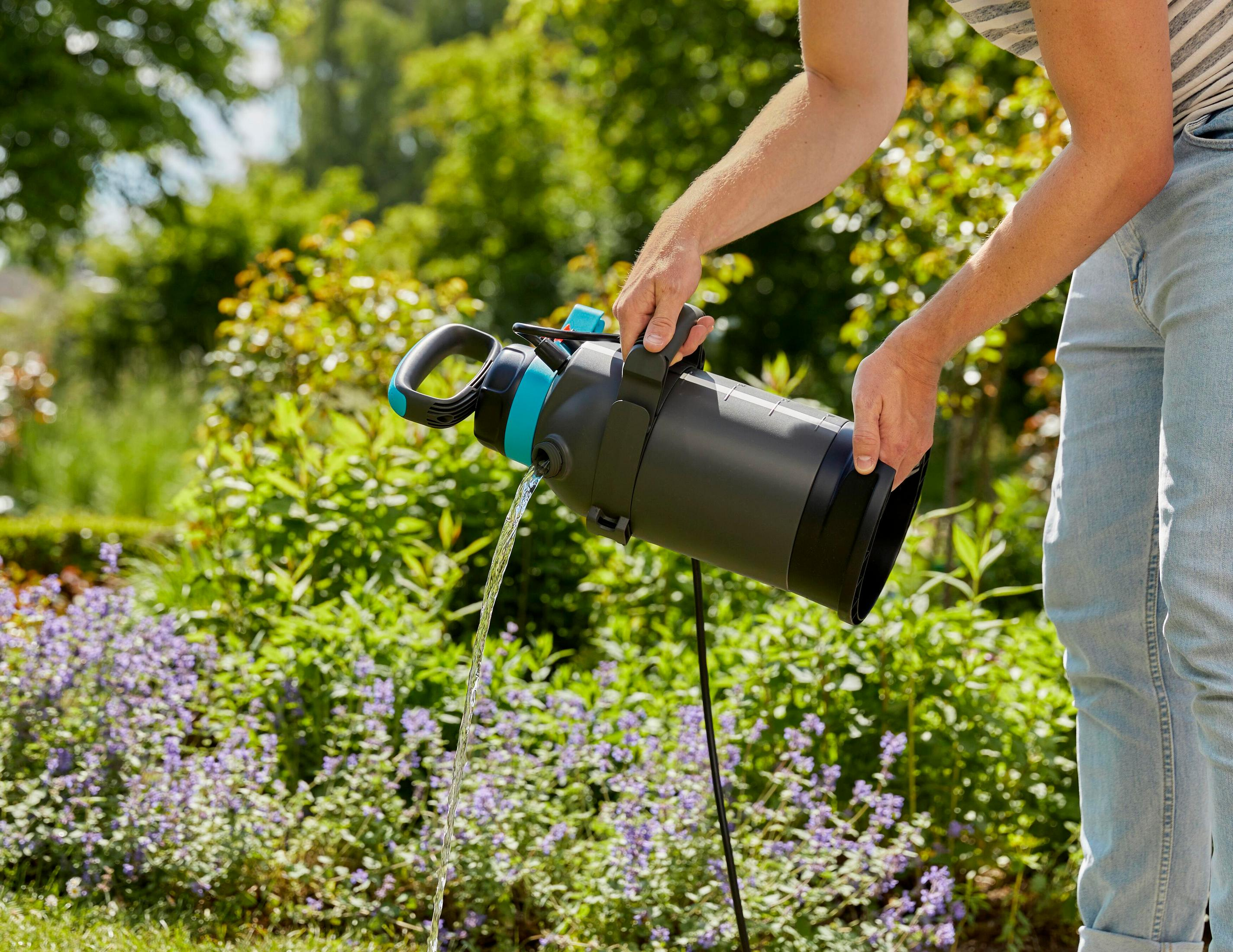 Person gießt Wasser aus schwarzem Behälter mit blauen Akzenten in einen Garten.