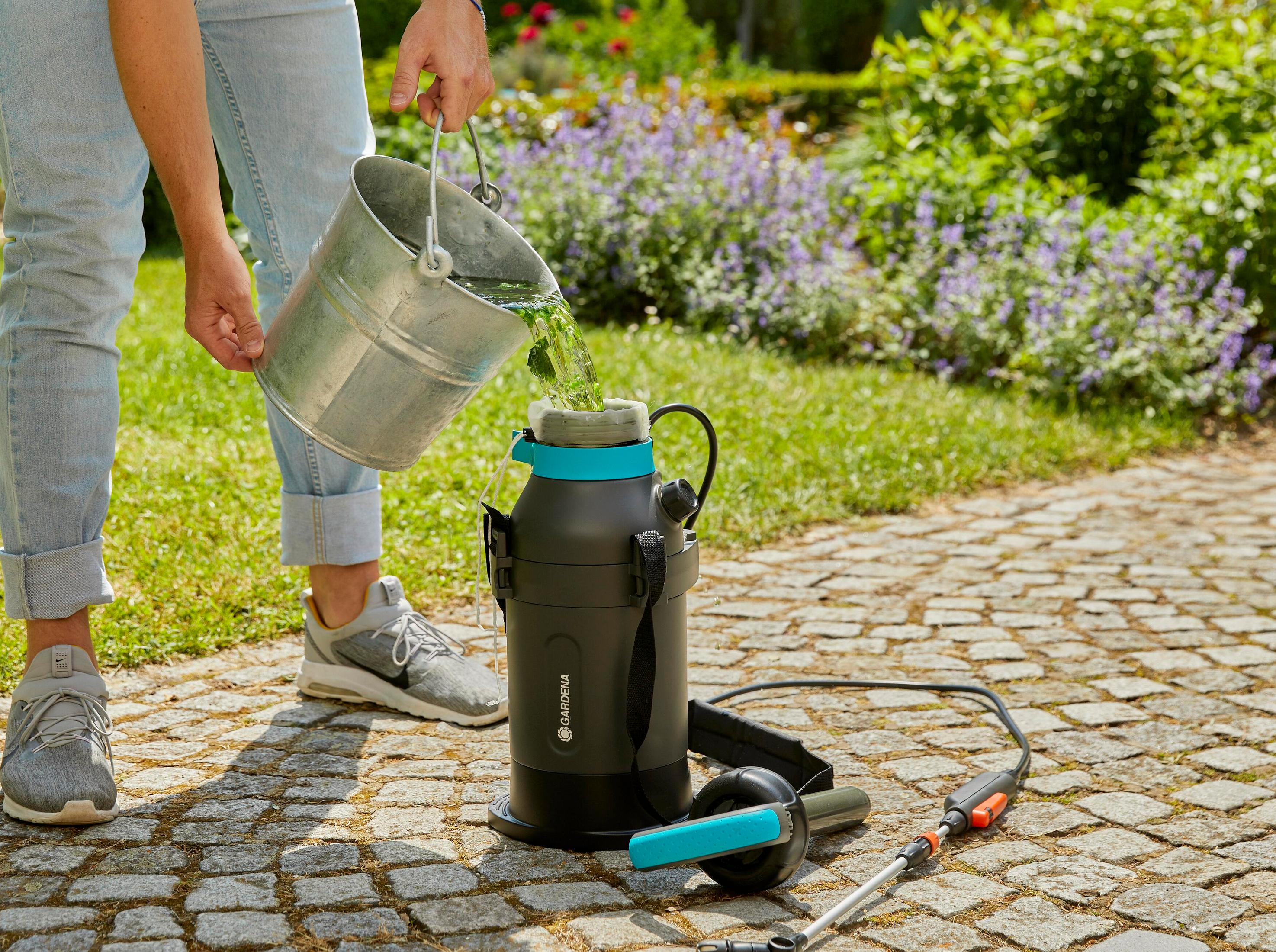 Person füllt Flüssigkeit aus Eimer in Sprühgerät auf gepflastertem Weg im Garten.