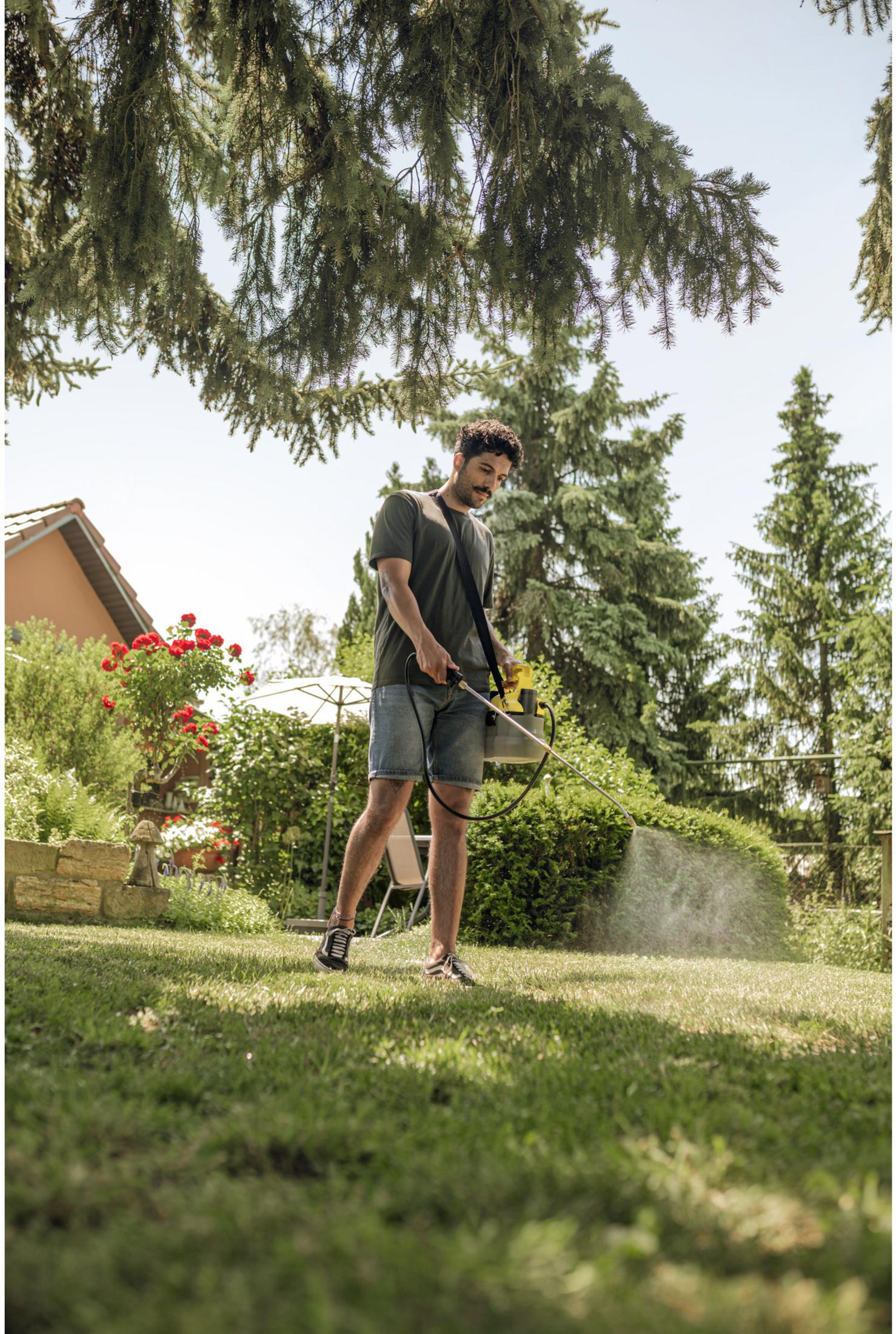 Mann sprüht Rasen mit gelbem Gerät in einem Garten mit Bäumen und Haus.