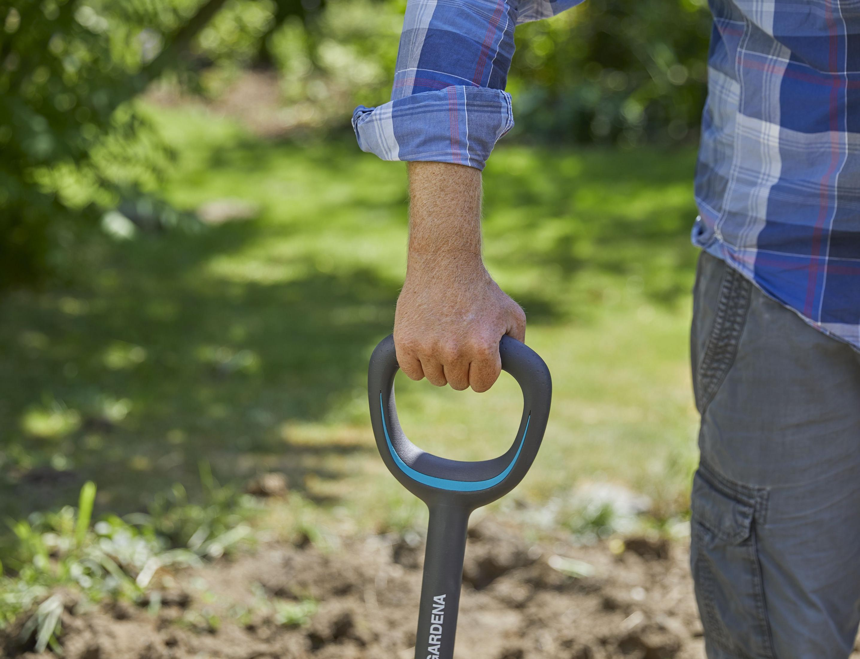 Uomo tiene manico pala, indossa camicia a quadri e pantaloni cargo in giardino.