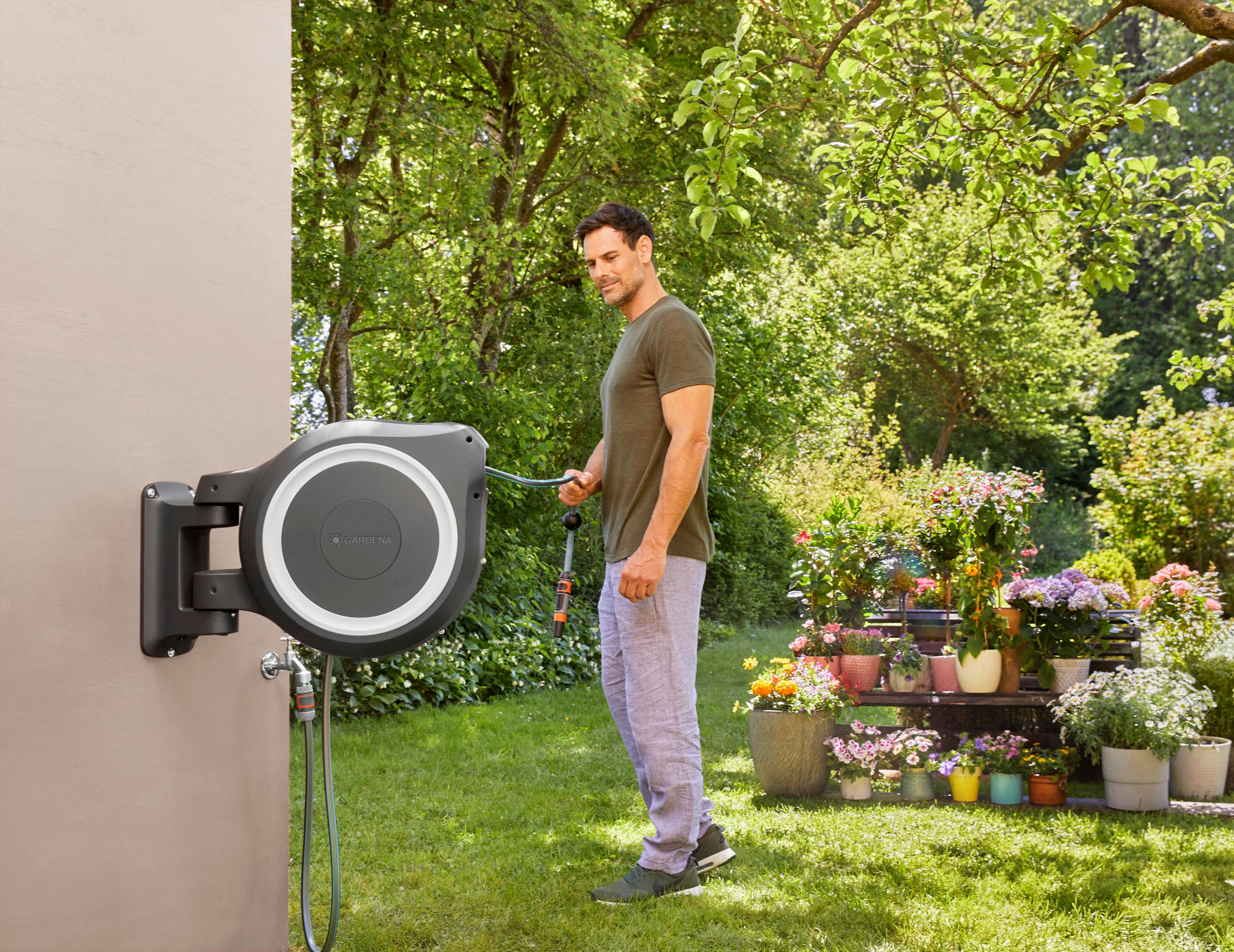 Mann mit Gartenschlauch neben Wandmontage-Schlauchaufroller und Blumen.
