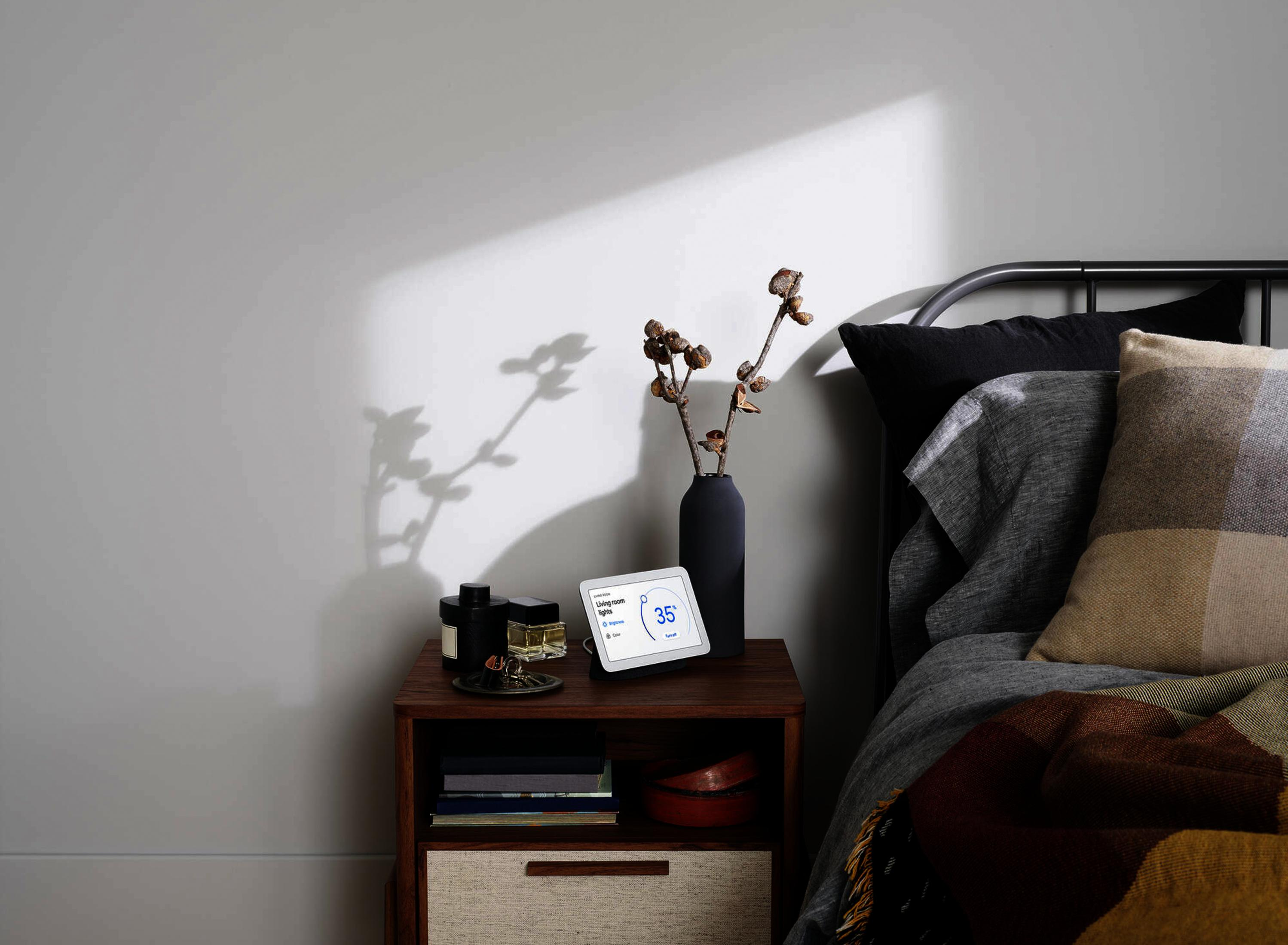 Una mesita de noche junto a una cama, con pantalla inteligente, jarrón con ramas y libros.