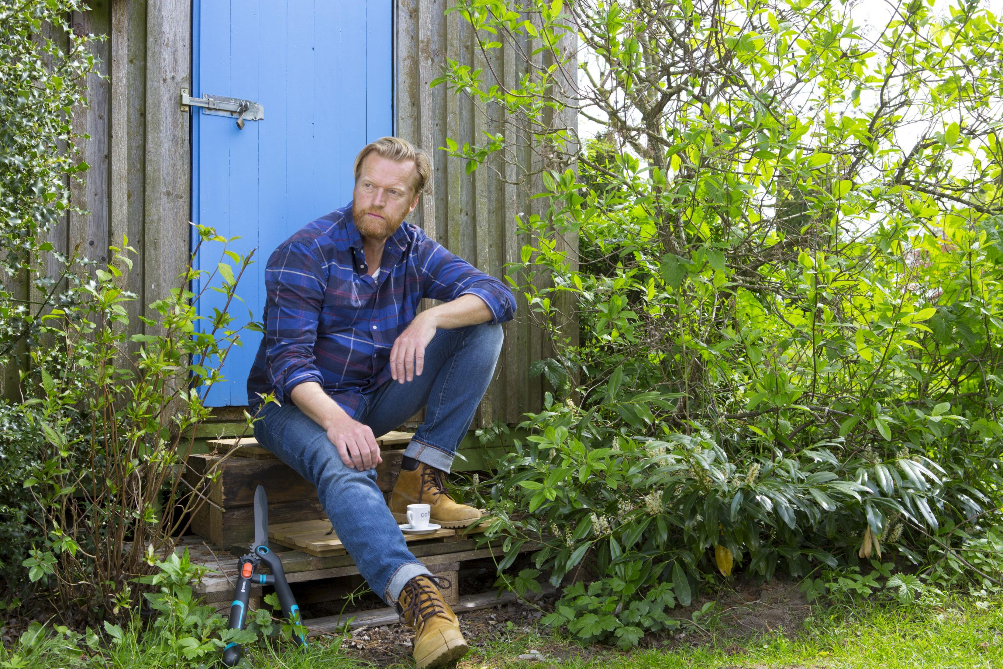Mężczyzna siedzi na drewnianych schodkach przed niebieskimi drzwiami obok krzaków.