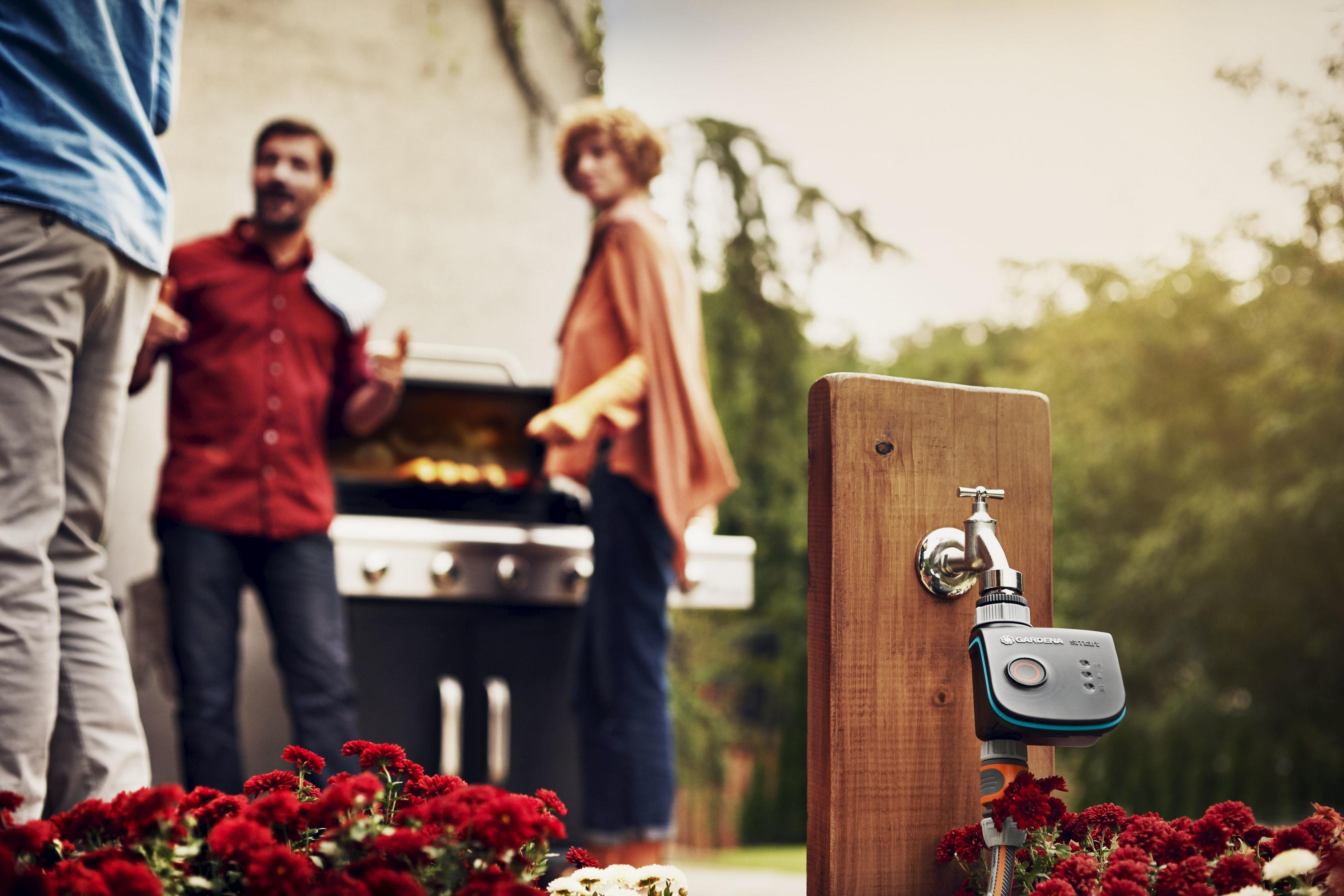 Un rubinetto con un controller di irrigazione intelligente su un palo di legno, con un barbecue sullo sfondo.