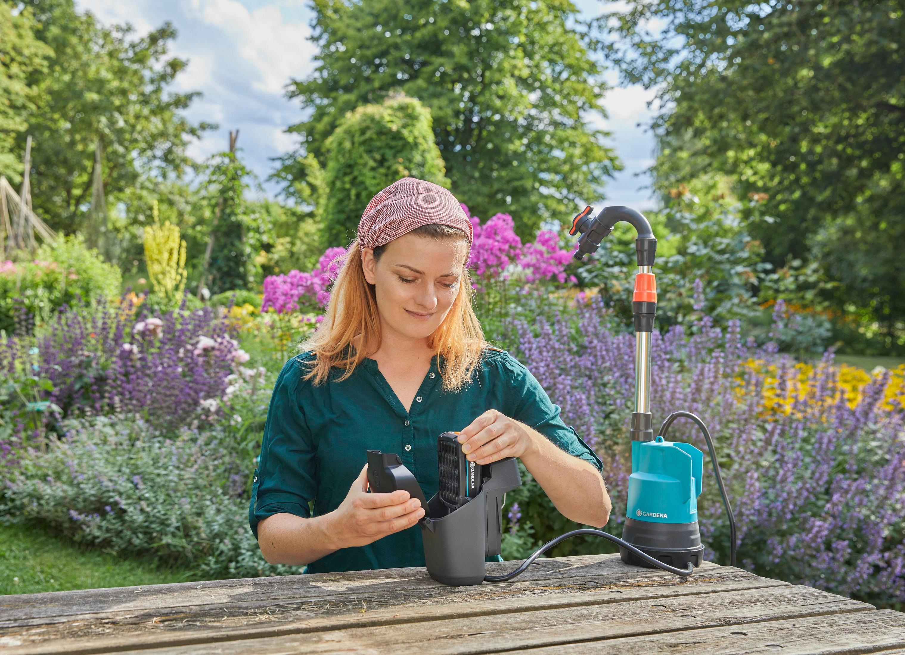 Une femme avec un foulard insère une batterie dans un appareil, dans un jardin.
