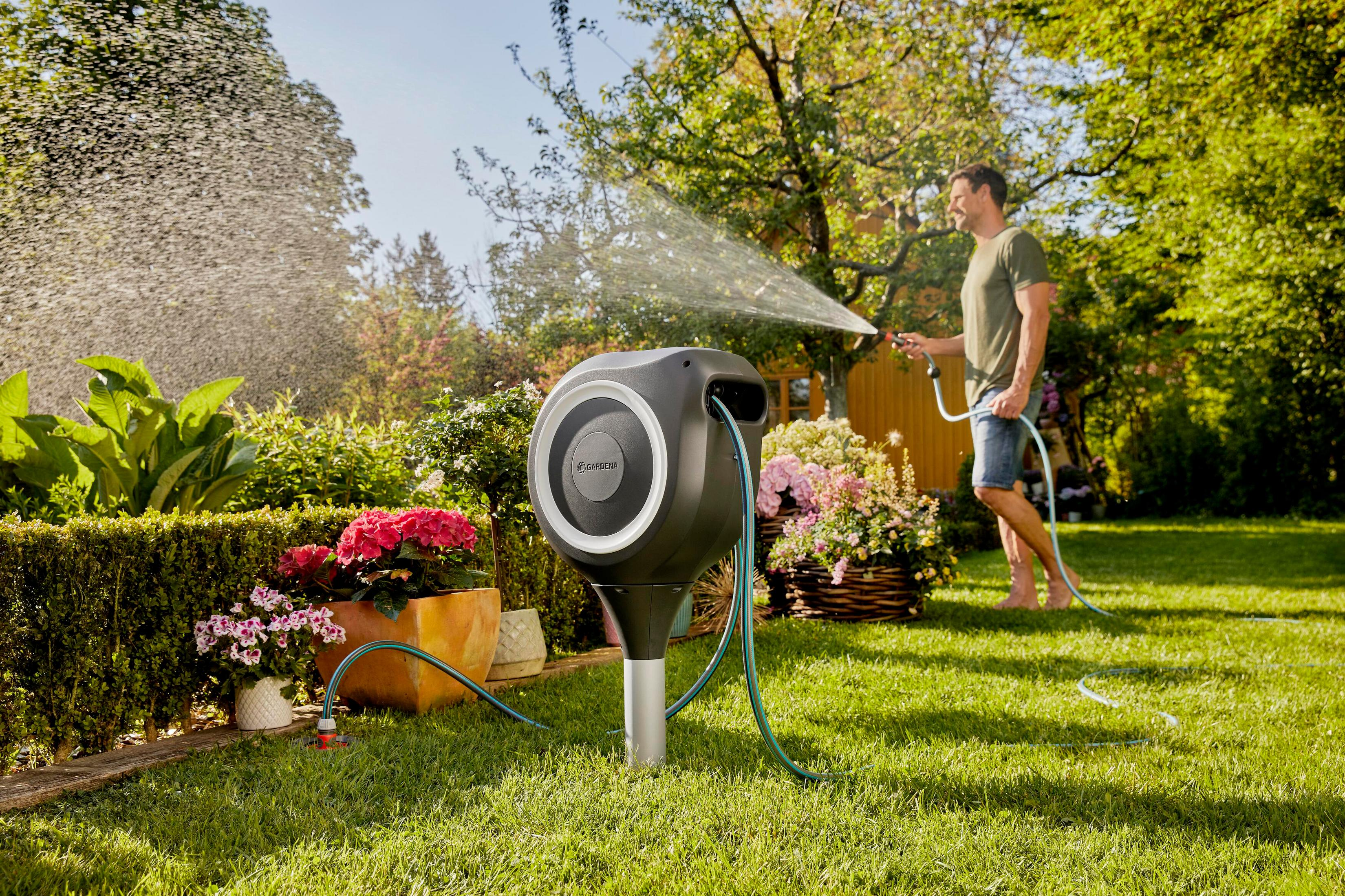 Uomo innaffia giardino con tubo, avvolgitubo in primo piano.