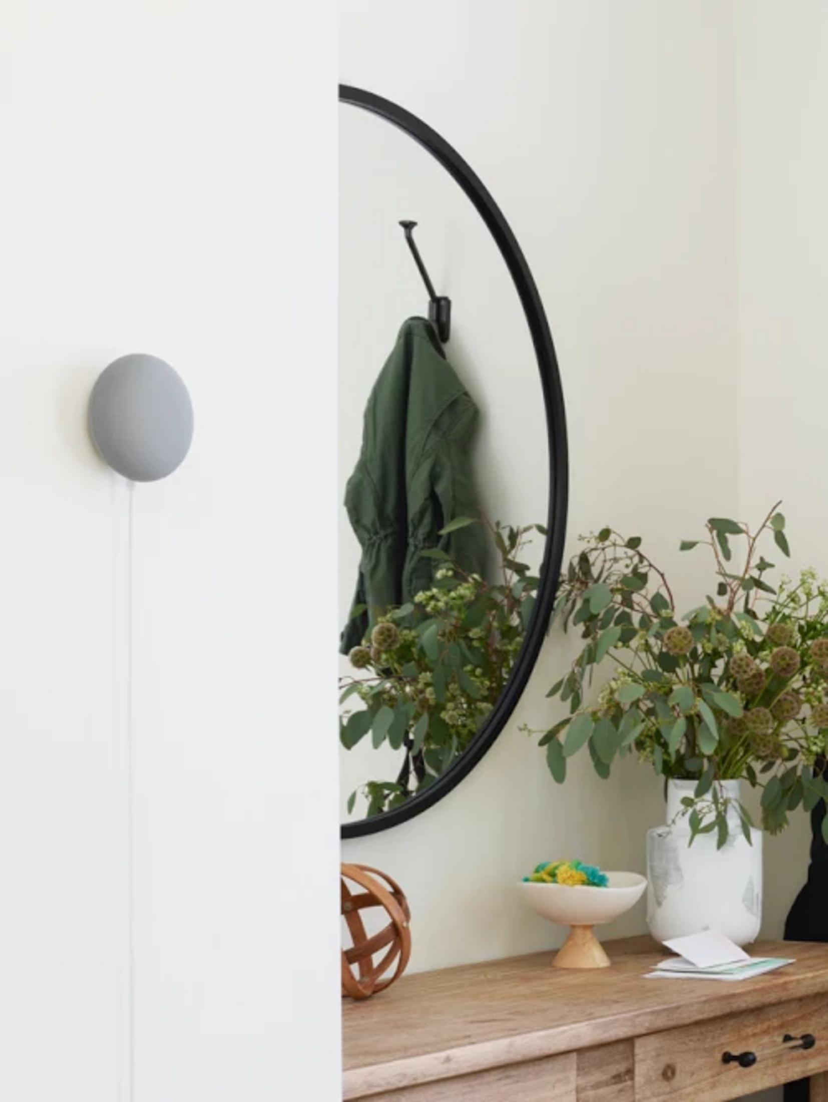 Okrągłe lustro z zielonym płaszczem na haczyku. Przed nim stół z wazonem i kwiatami.