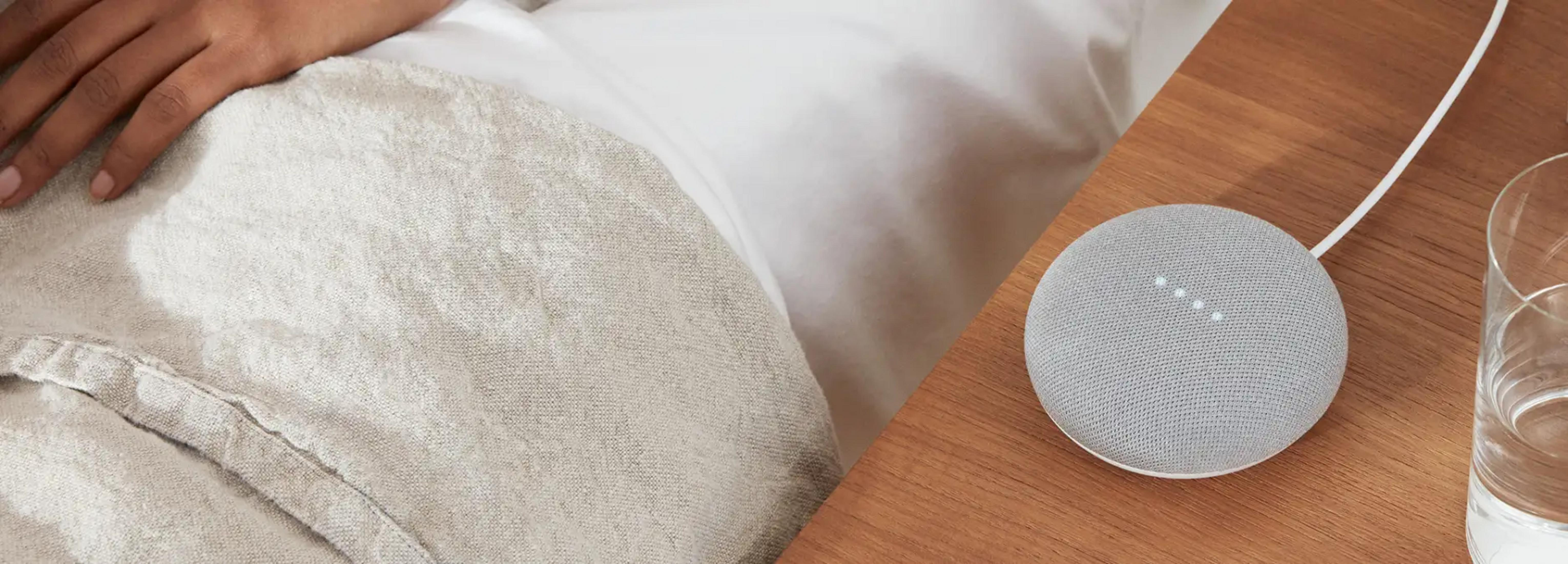 Osoba odpoczywa w łóżku obok inteligentnego głośnika na szafce nocnej.