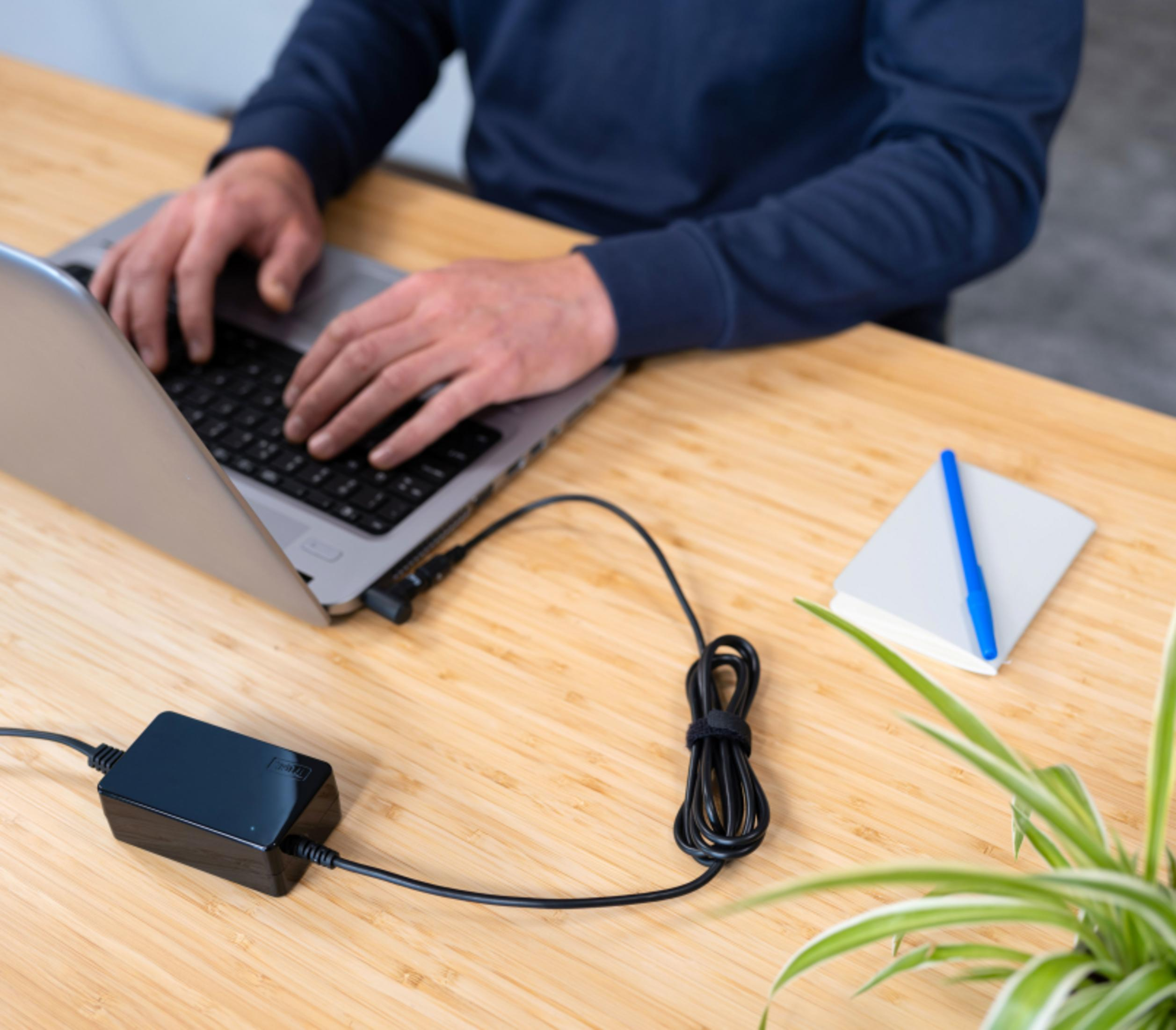 Persona escribe en portátil, adaptador de corriente y cuaderno en escritorio.