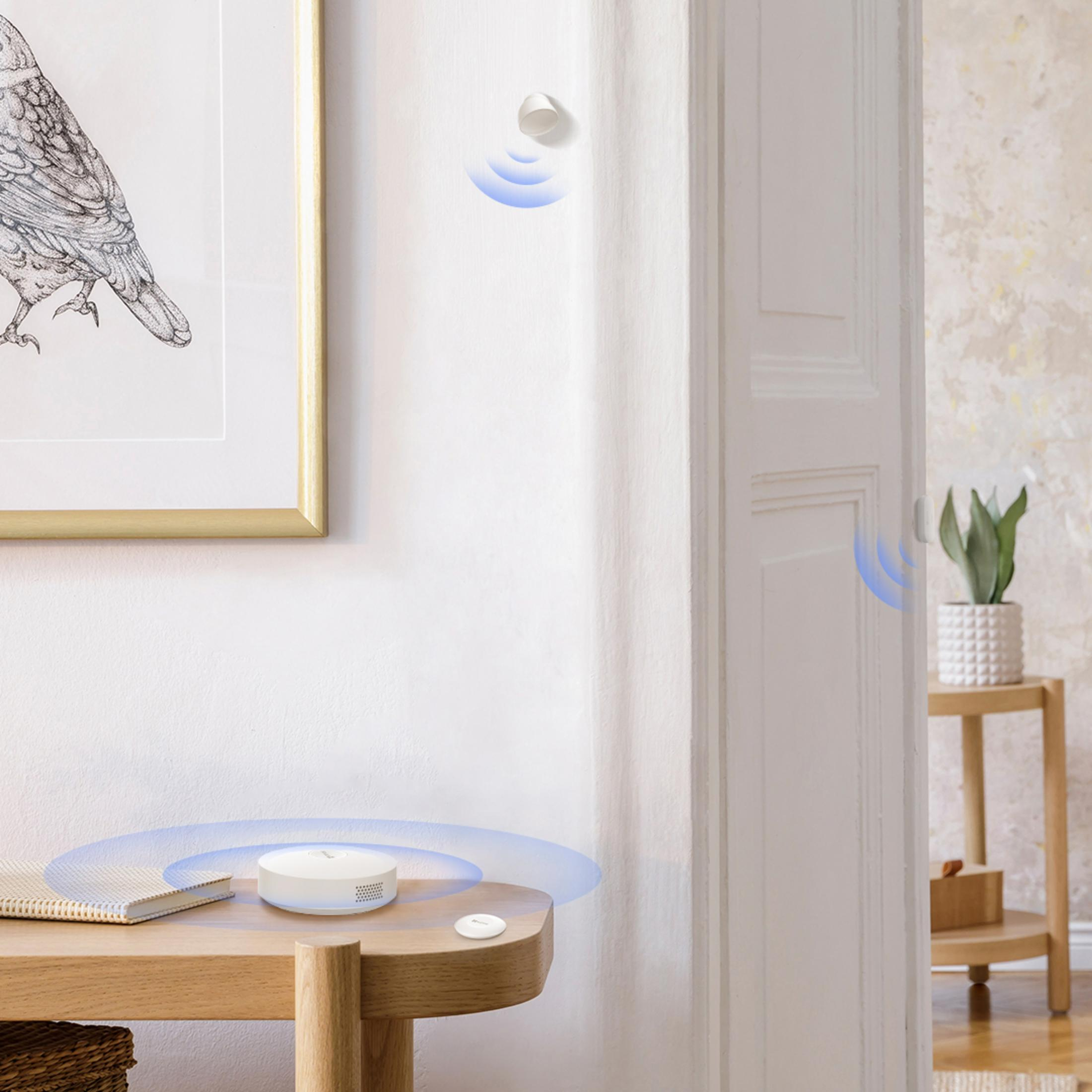 Biały czujnik smart home na drewnianym stole obok książki.