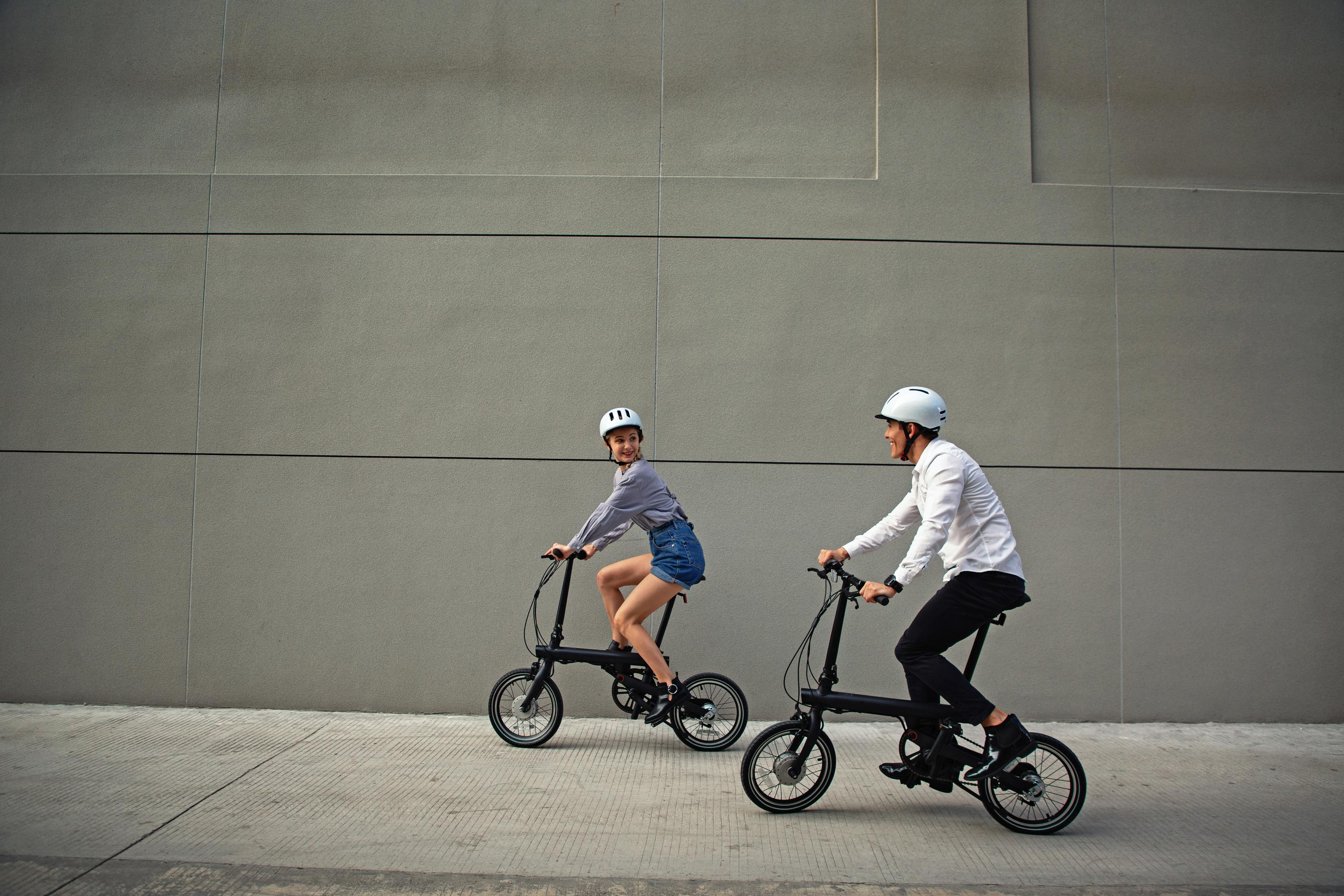 Due persone su biciclette pieghevoli pedalano davanti a un muro grigio.
