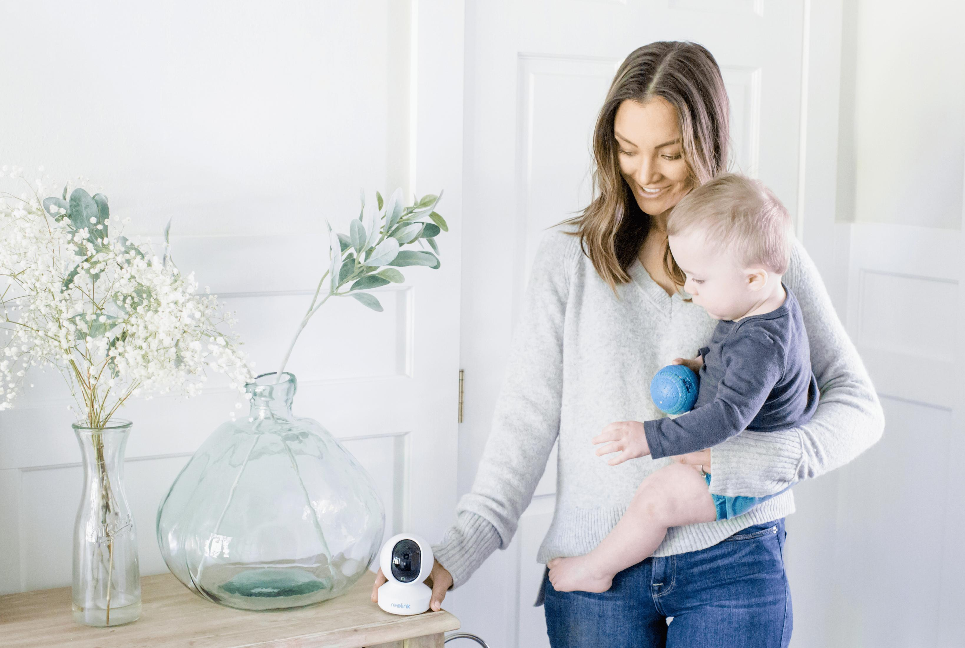 Eine Frau hält ein Baby. Auf einem Tisch stehen Blumen und eine Kamera.