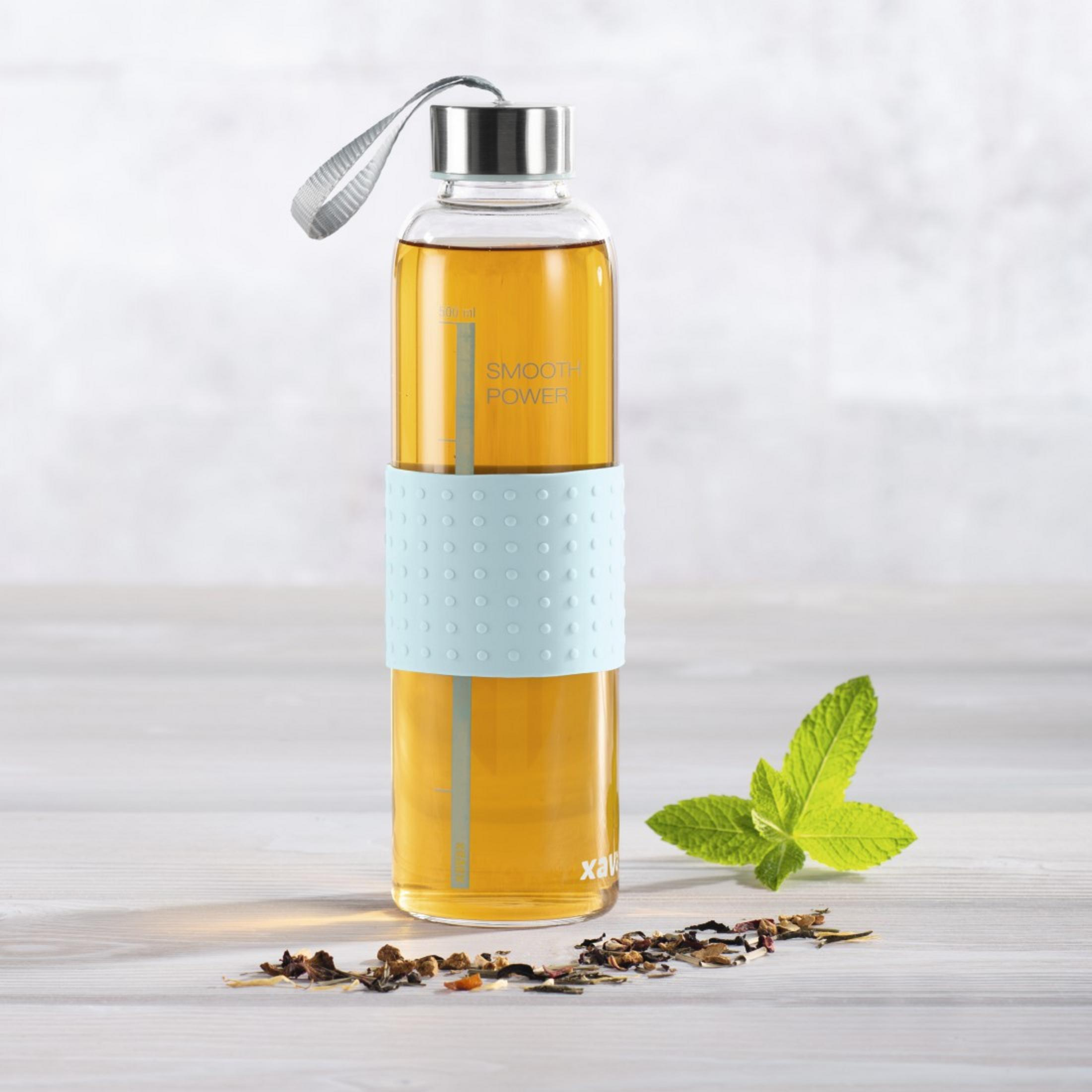 Glasflasche mit Tee, Minzblatt und Teeblättern auf hellem Holztisch.