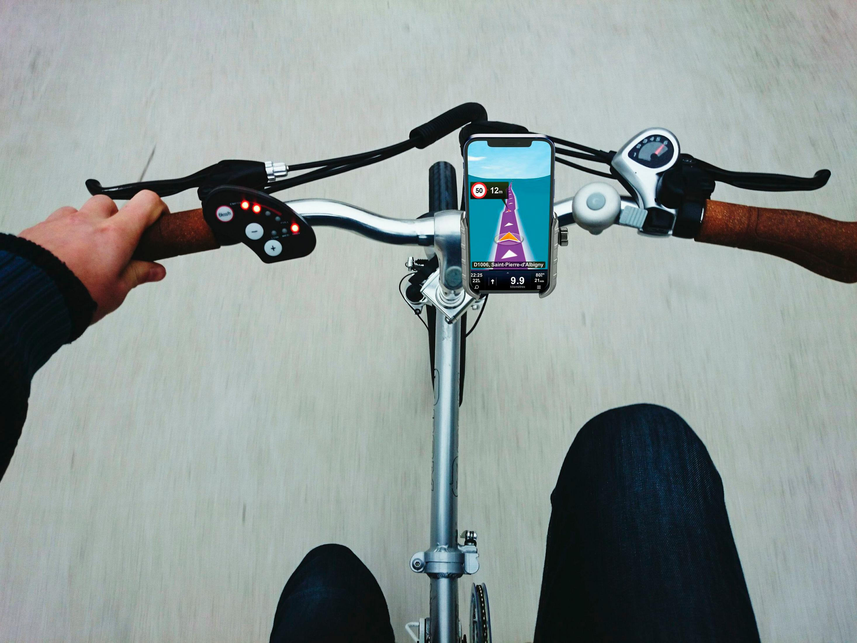 Vista in prima persona in bicicletta con telefono che mostra navigazione GPS. Volante, controlli e gambe visibili.